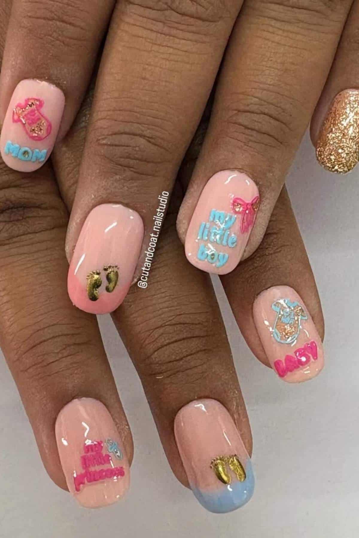 A hand with nude pink nails features colorful 3D stickers with words like "MOM," "BABY," "LITTLE BOY," and small shoe designs; one nail is painted with gold glitter.