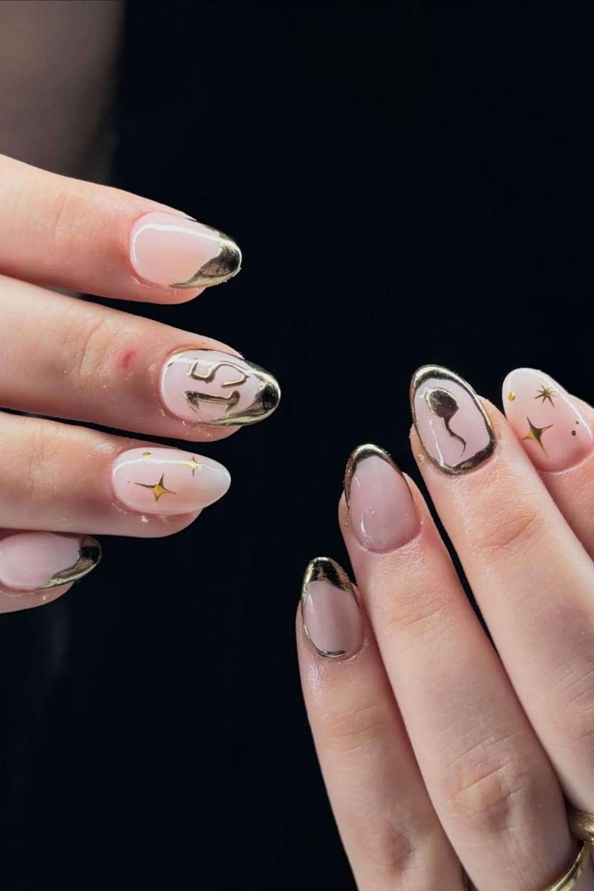Close-up of hands with almond-shaped nails featuring nude polish, gold abstract lines, stars, and metallic gold tips in a minimalistic nail art design.