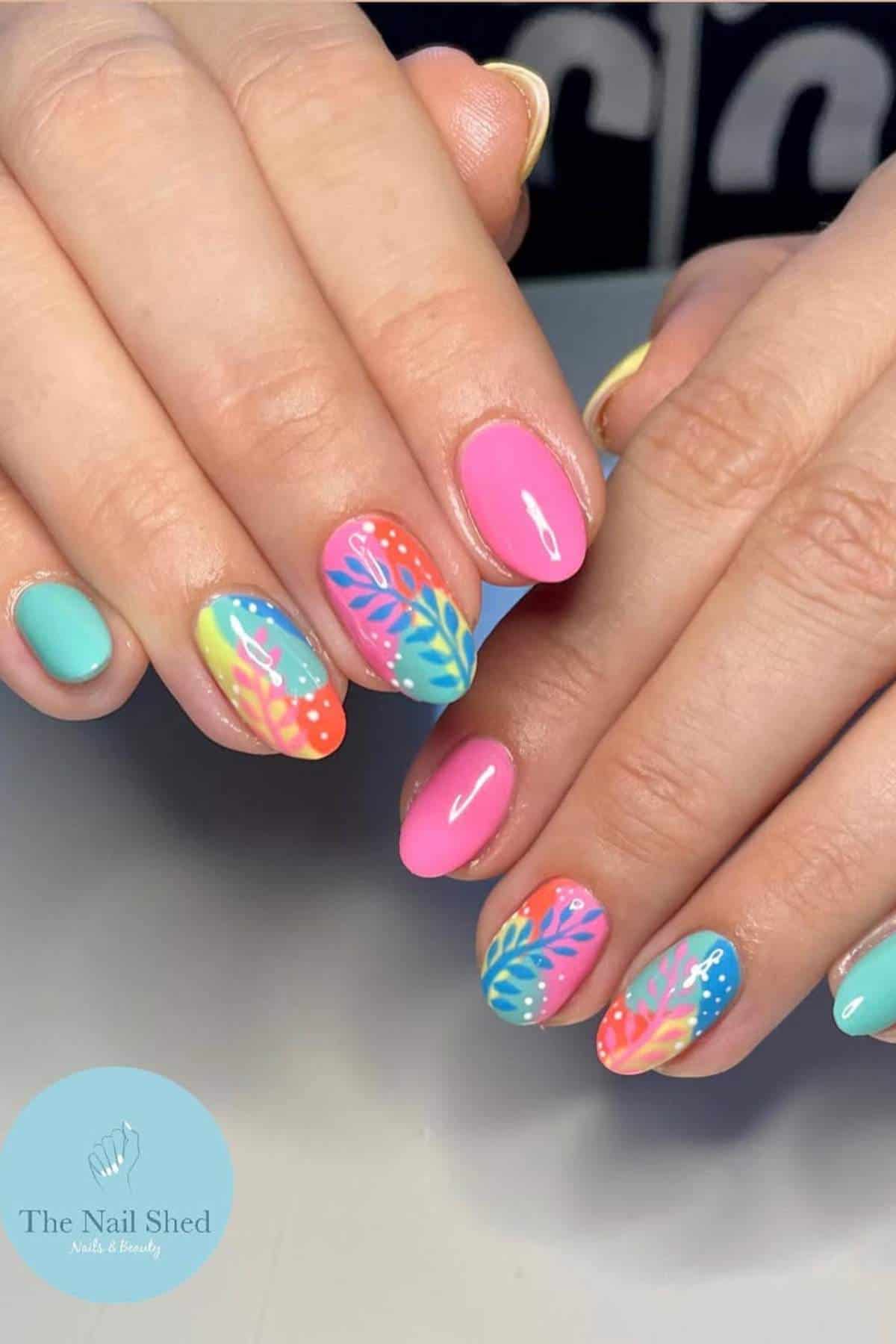Hands with brightly colored, oval-shaped nails featuring pink, blue, and turquoise polish, some with intricate multicolored leaf patterns. Nail salon logo visible in the corner.