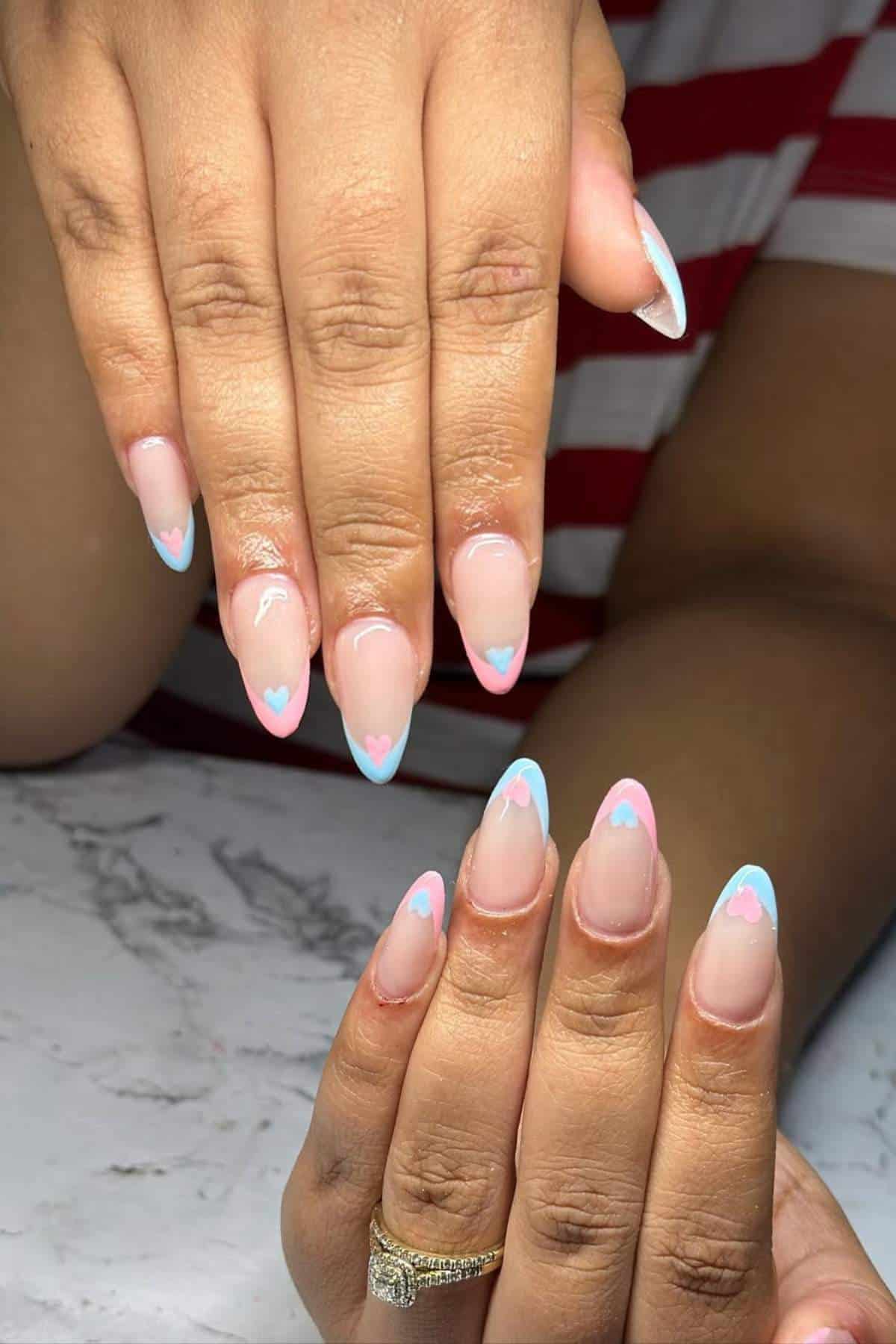 Hands with almond-shaped nails featuring a nude base, blue French tips, and small pink heart designs at the tips; a gold ring is worn on one finger.