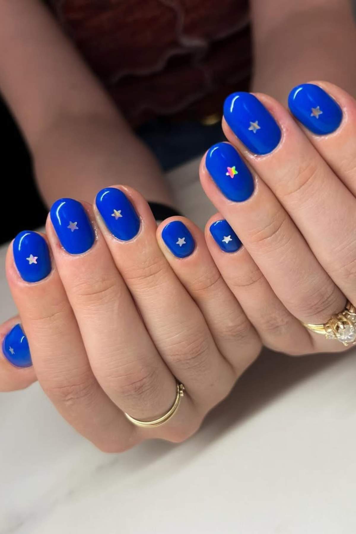 Hands with short, oval-shaped nails painted bright blue, each nail decorated with a small silver star sticker; one nail has a colorful star.