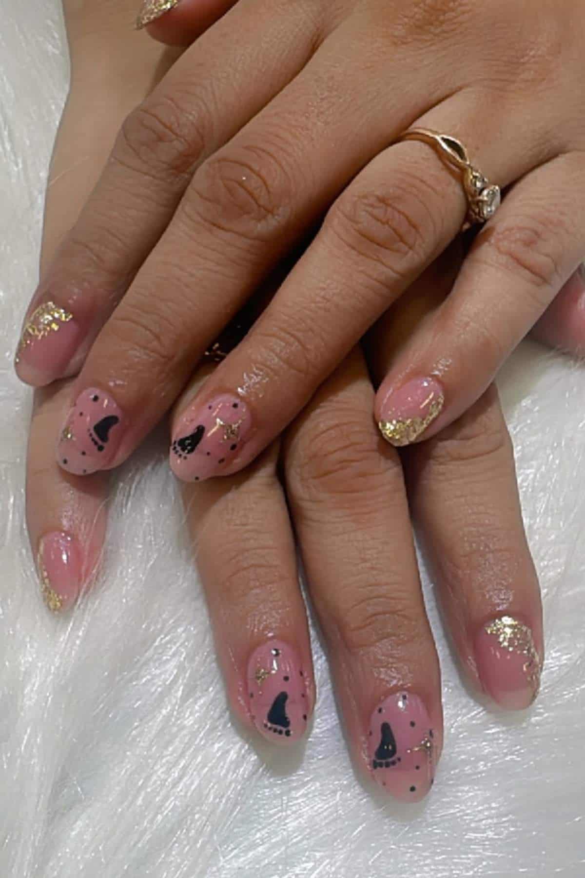 Two hands with manicured nails featuring pink polish, black abstract designs, and gold glitter tips, resting on a white textured surface.