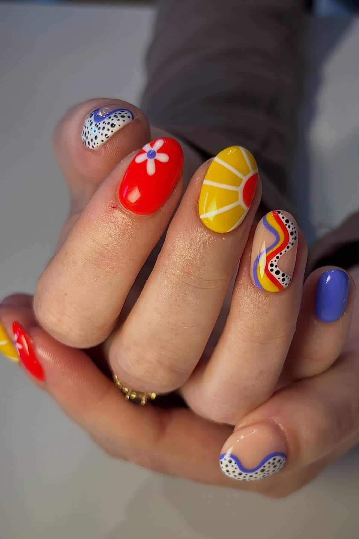A hand with colorful, patterned nail art including florals, geometric shapes, wavy lines, dots, and solid colors in red, yellow, blue, purple, white, and black.