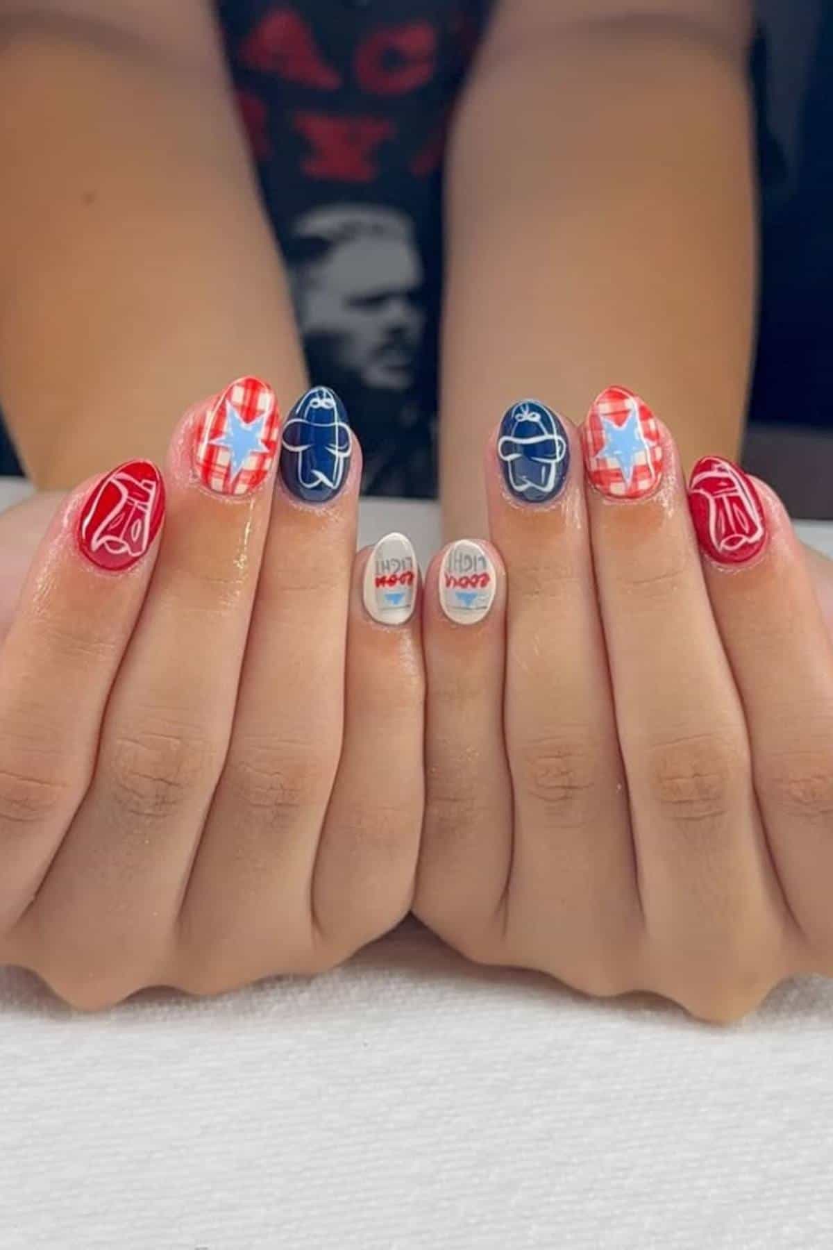 Close-up of hands with red, white, and blue nail art featuring abstract patterns, checkered designs, and text reading "Hot Yachty" on the middle fingernails.