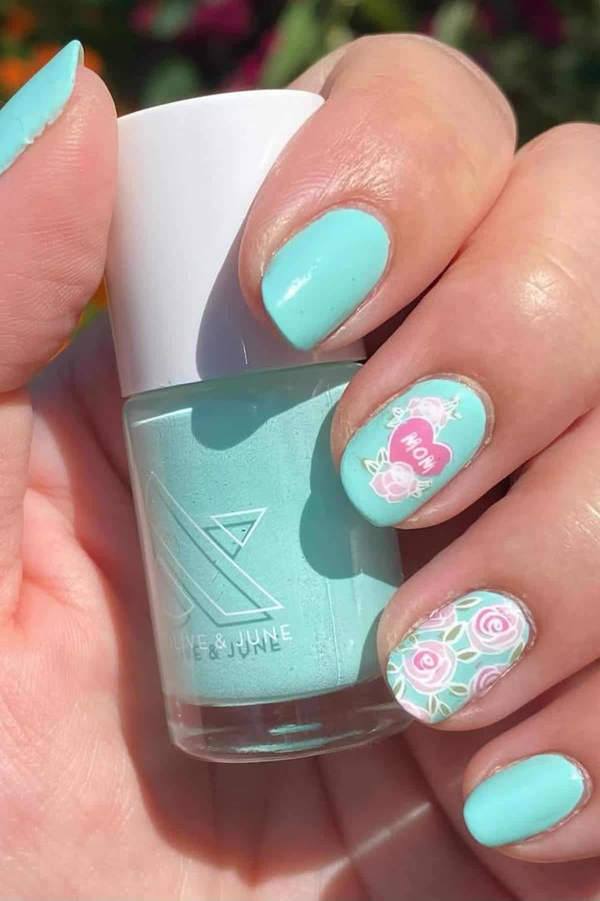 A hand holding a bottle of mint green nail polish with manicured nails featuring mint green color and floral and "Mom" designs on accent nails.
