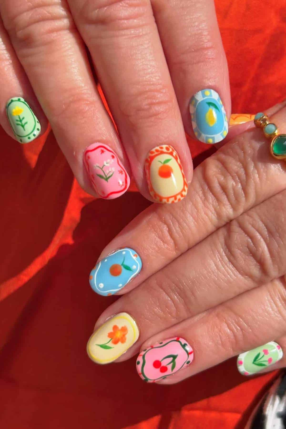 Close-up of hands with colorful, hand-painted nail art featuring various floral and fruit designs on each fingernail.