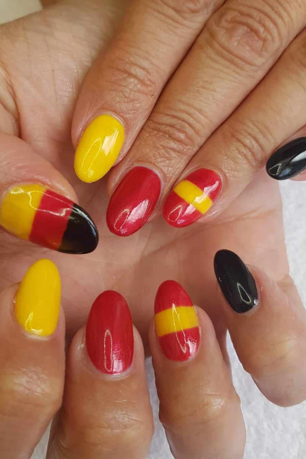 Hands with manicured nails painted in red, yellow, and black with various geometric designs, including stripes and color blocking, on each fingernail.
