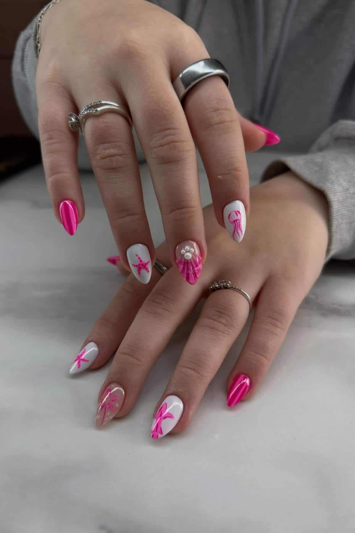 Hands with long, almond-shaped nails painted pink and white, featuring sea star, seashell, and bow designs, accented with small pearls. Person wears silver rings.