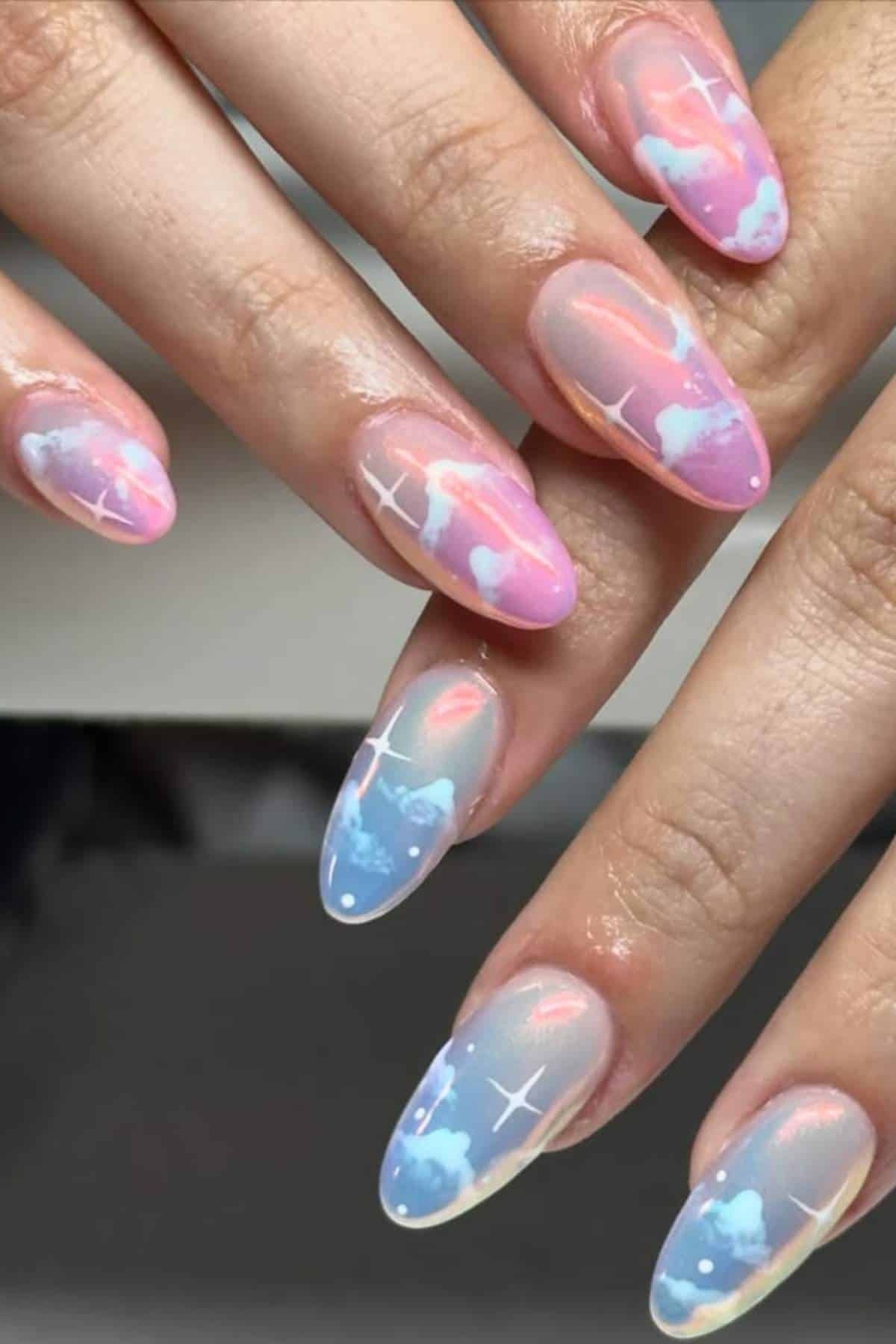 Hands with long, almond-shaped nails featuring a pastel gradient design in pink, blue, and white, decorated with clouds and small white star accents.