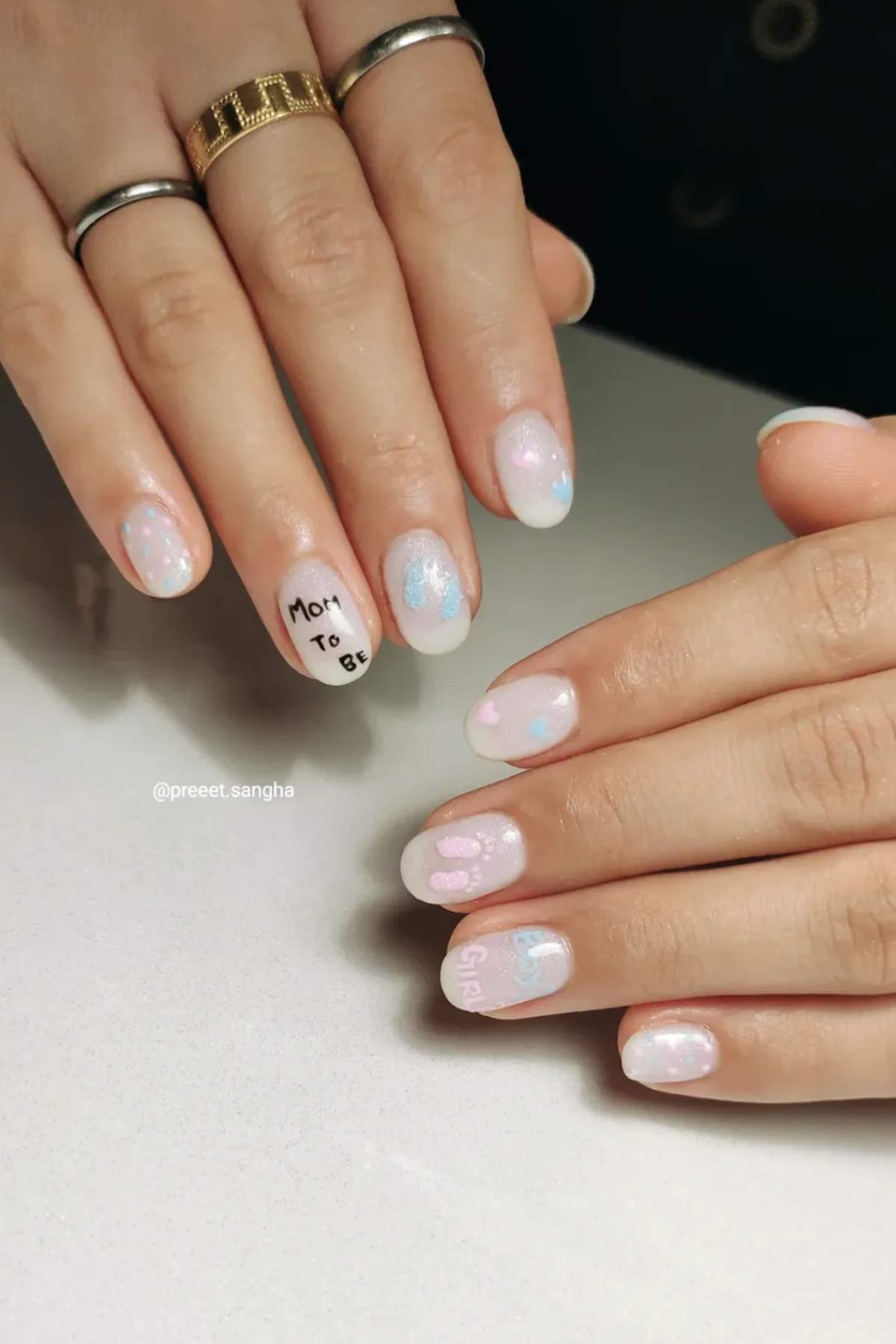 Close-up of hands with pastel pink and blue gradient nail art; one nail features the text "Mom to be" and others have small heart and dot designs.