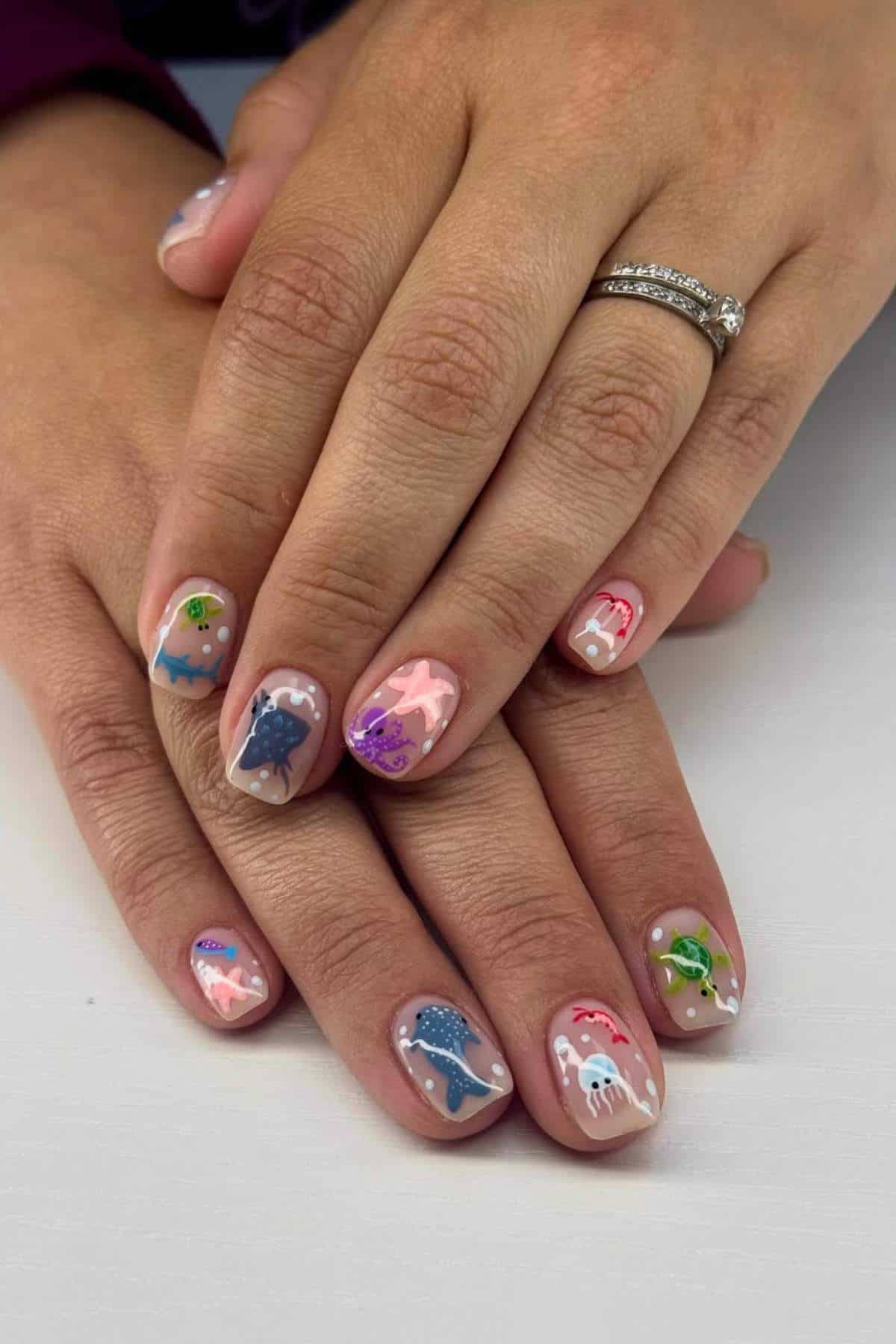 Hands with short nails painted with colorful sea creature designs, including fish, jellyfish, and starfish, on a light nude base. The person wears a silver ring.