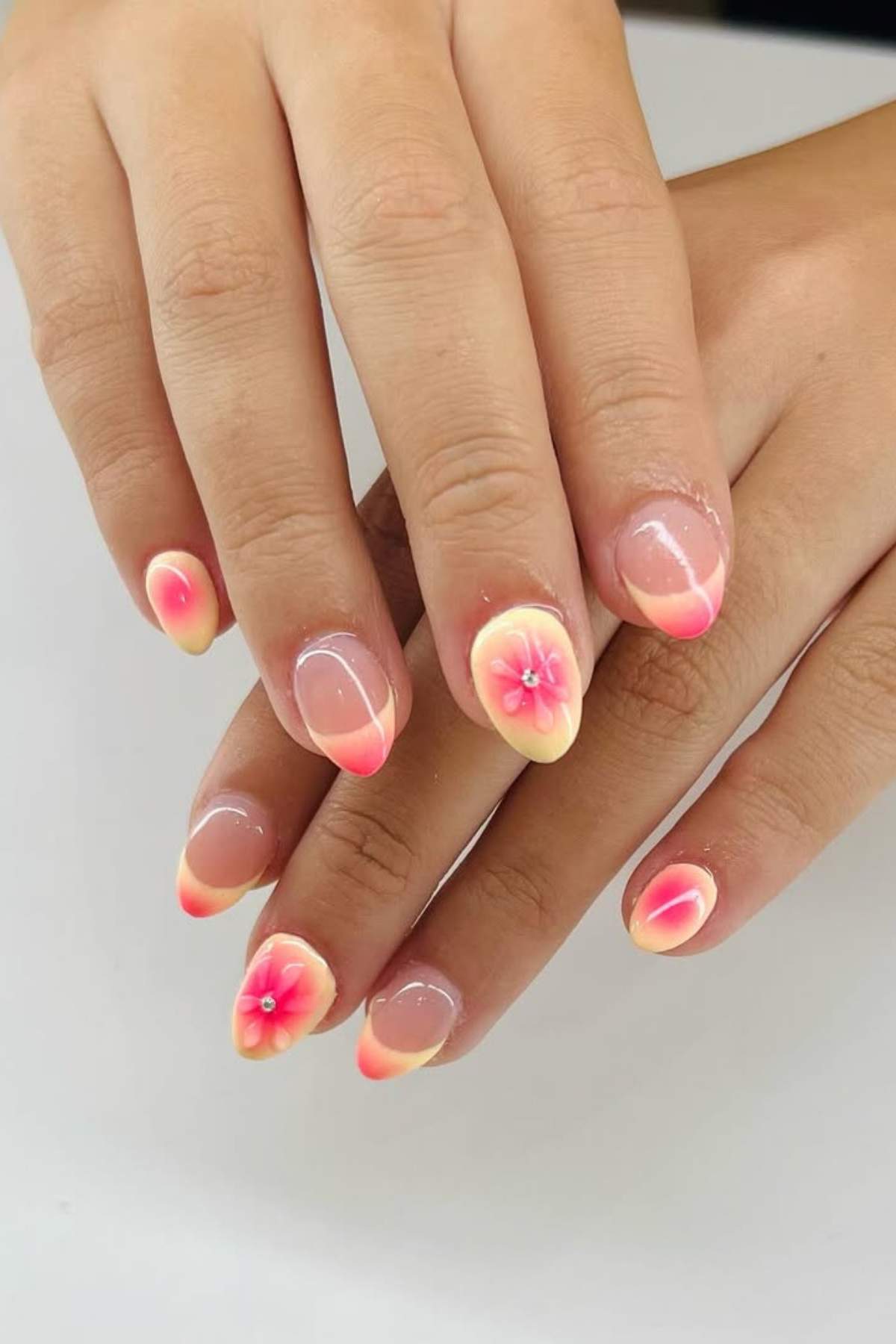 Hands with manicured nails featuring a pink and yellow gradient and 3D floral designs on each nail, set against a plain background.