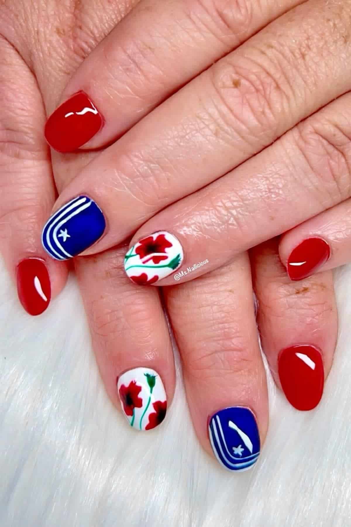 Hands with red, blue, and white nail art; some nails have solid colors, others feature flowers and a striped blue design with a white star.