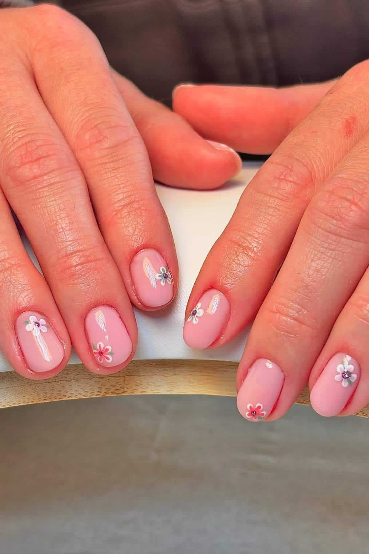 Hands with short, rounded nails painted light pink, each nail decorated with a small, white and pink flower design.