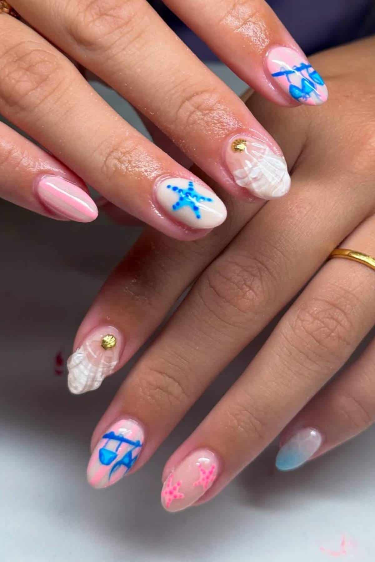 Close-up of hands with manicured nails featuring ocean-themed designs, including shells, starfish, and waves in blue, pink, and white hues with gold accents.
