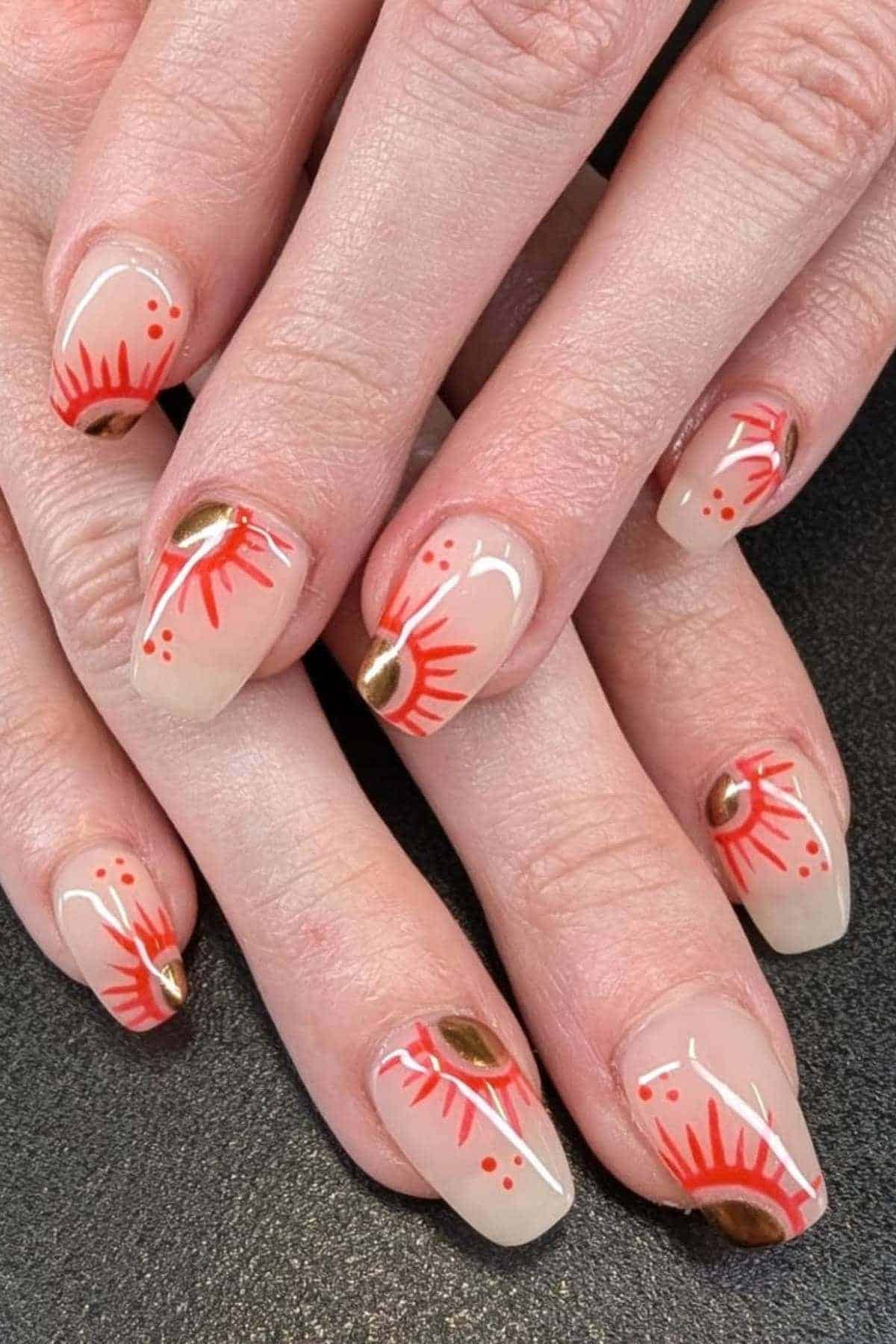 Fingernails with a beige base, decorated with red sunburst patterns and small gold oval accents on each nail.