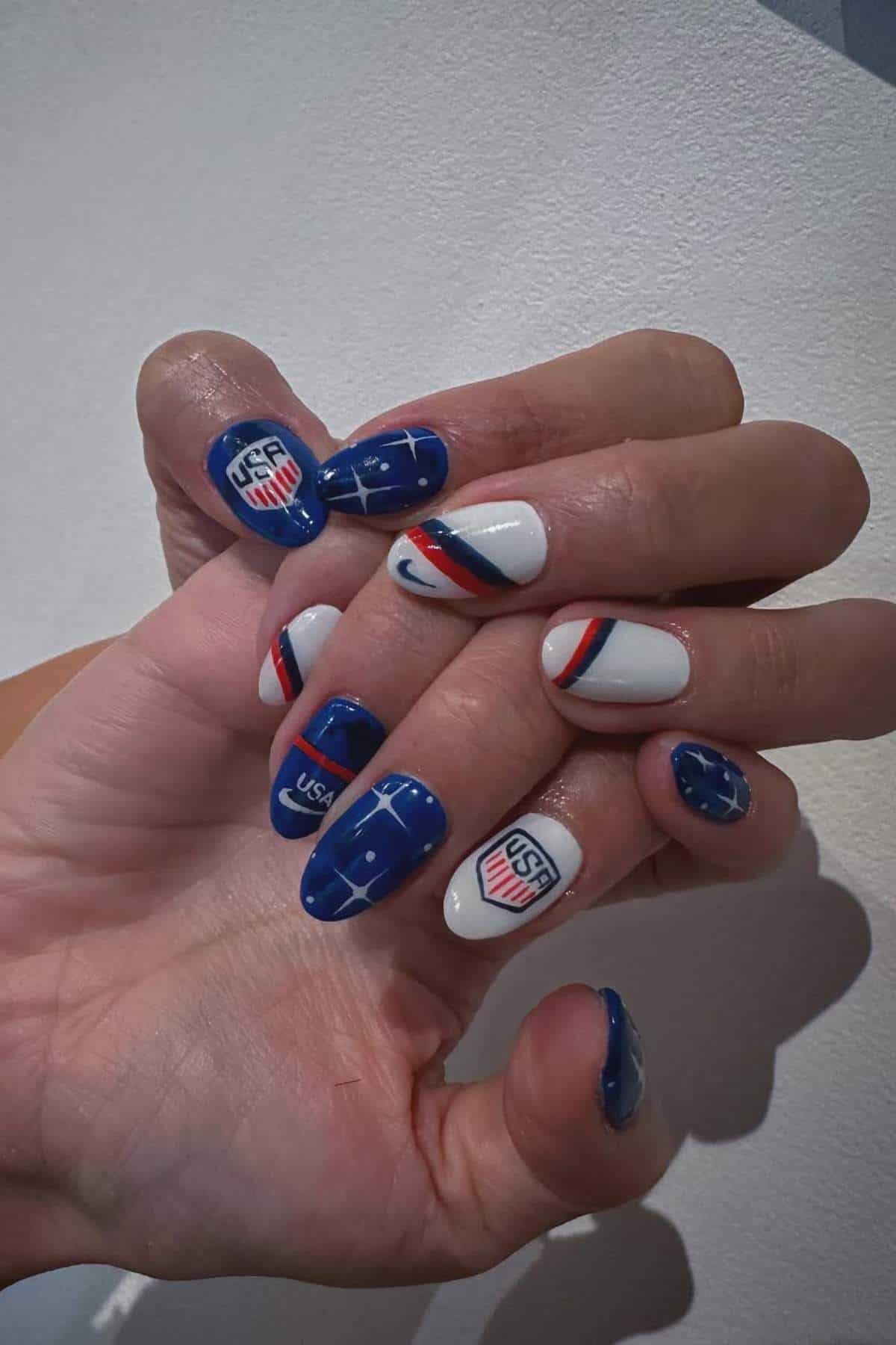 Close-up of hands with manicured nails featuring blue, white, and red designs, including the USWNT logo and soccer field patterns.