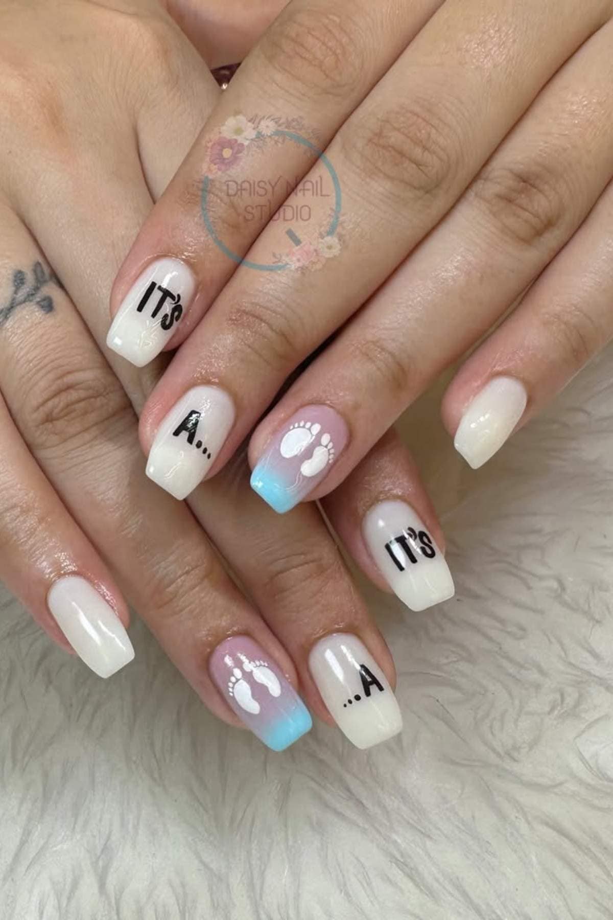 Close-up of hands with square-shaped nails; two nails have the text "It's a..." and two feature small white baby footprints on a light blue gradient background.