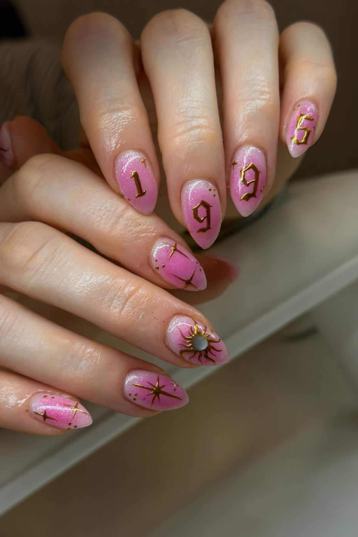 A hand with long, pointed pink nails featuring gold designs, including the numbers "1996," stars, and a sun motif.