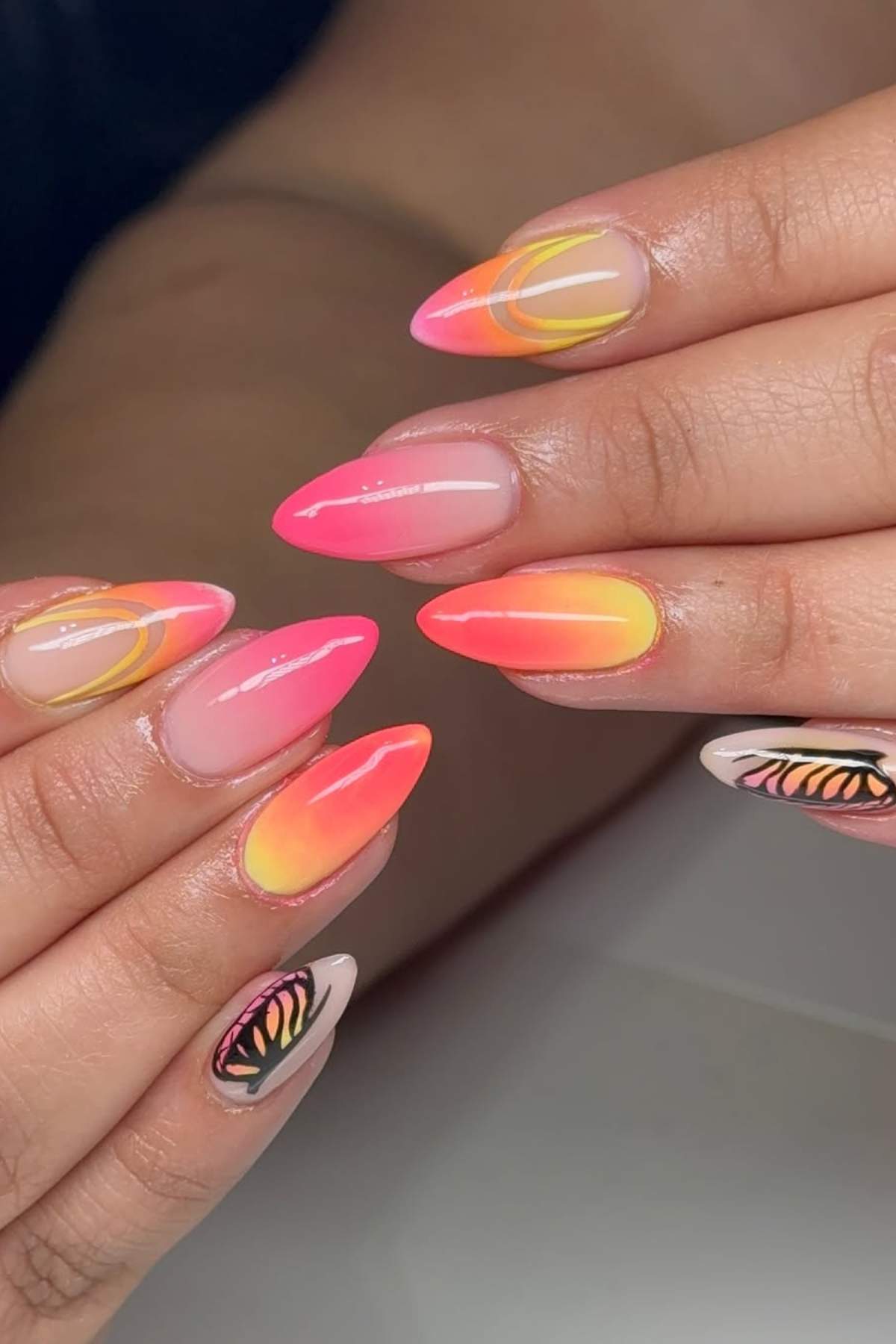 Hands with long almond-shaped nails featuring neon pink and orange ombre designs, abstract yellow lines, and butterfly wing patterns on the ring fingers.
