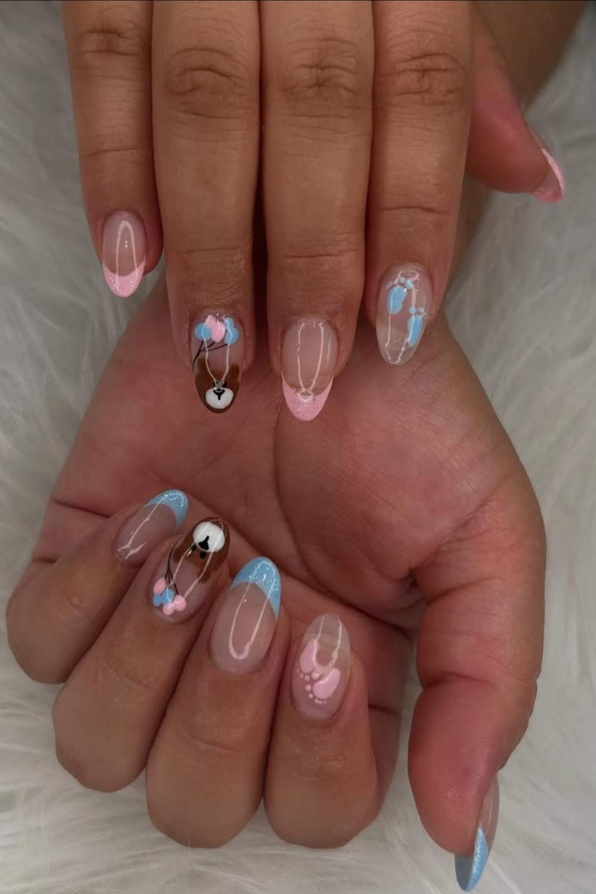 Hands with manicured nails featuring pink and blue French tips, heart designs, and balloon nail art on a soft white surface.