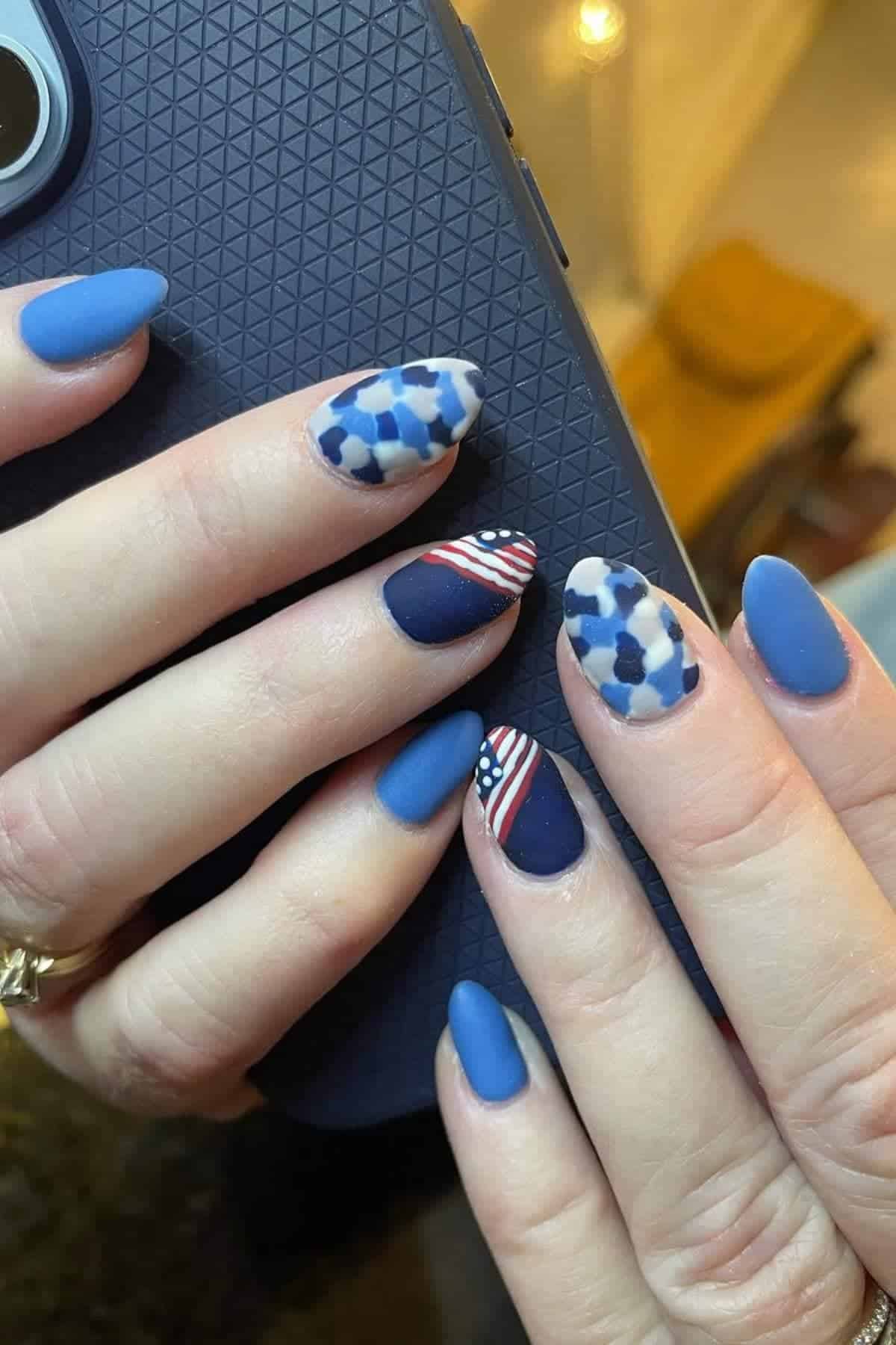 Hands with almond-shaped blue matte nails; some nails have a USA flag design and others have a blue and white geometric pattern, holding a textured dark phone case.