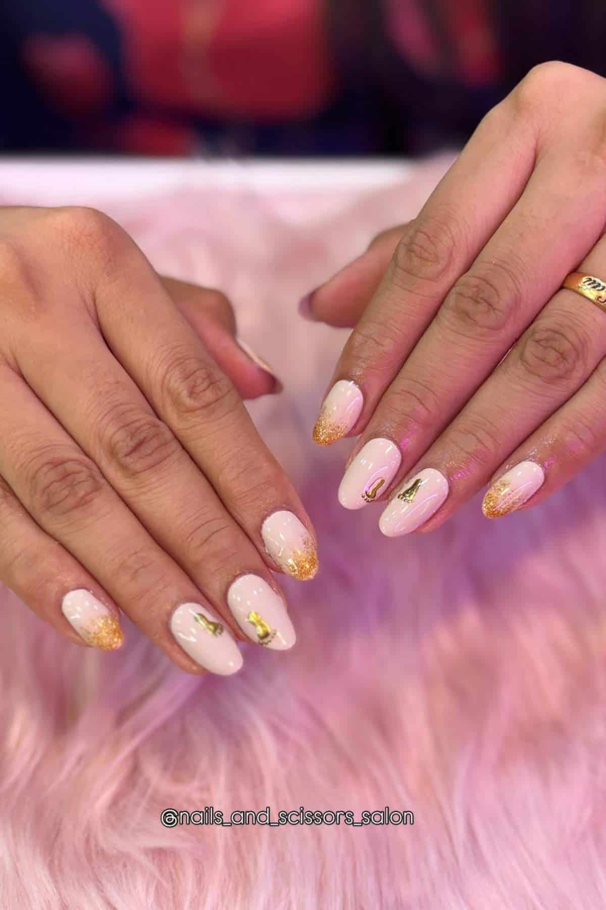 Hands with almond-shaped nails featuring a light pink base, gold foil accents, and glittery gold tips, shown resting on a pink furry surface.