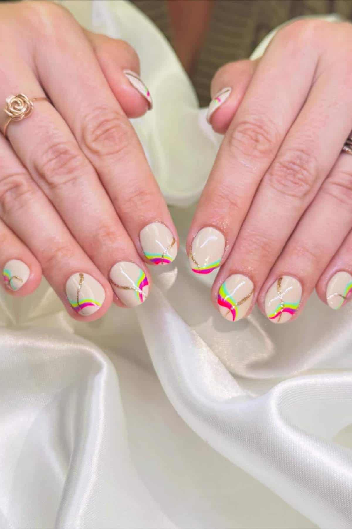 Hands with short, almond-shaped nails feature a nude base, colorful wavy lines in green, pink, and gold, and subtle gold accents; a rose-shaped ring is on one finger.