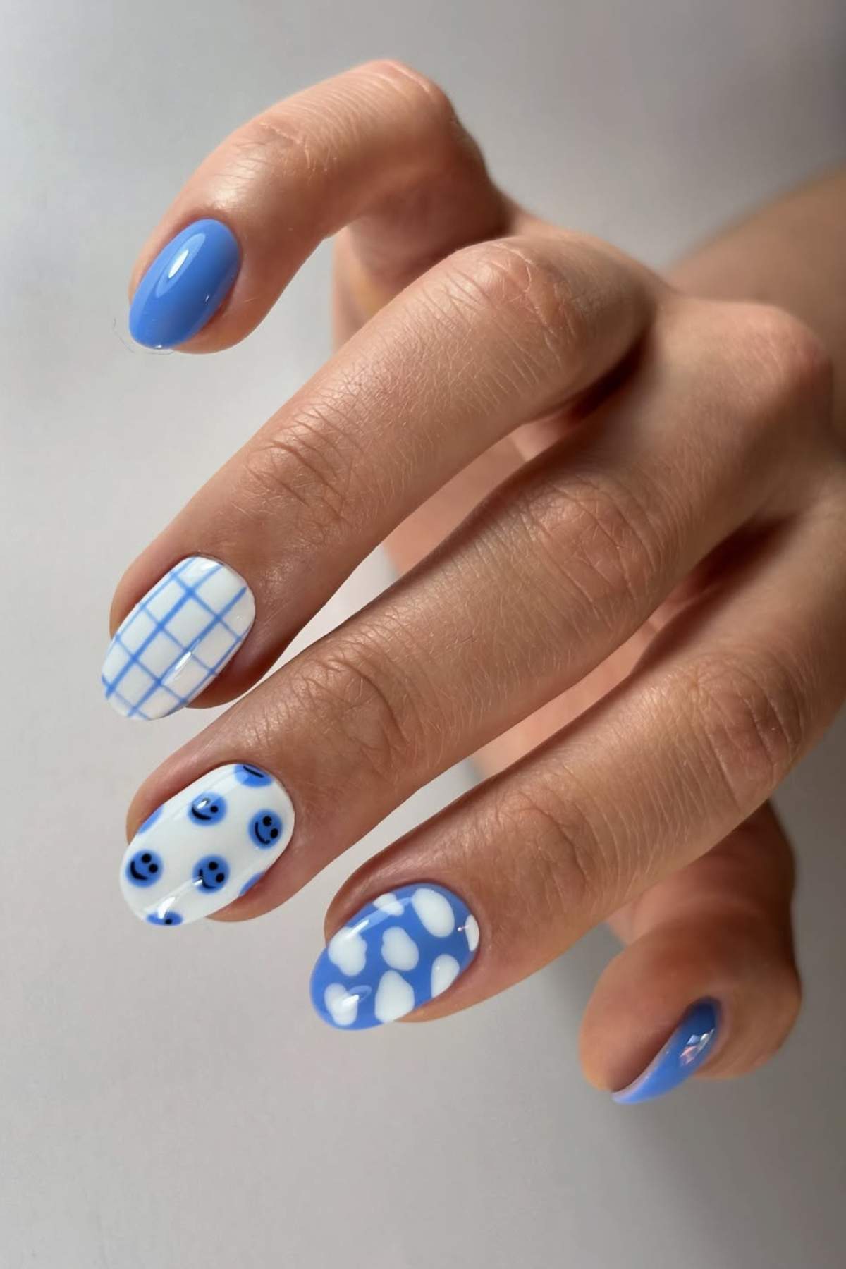 A hand with blue and white nail art featuring solid blue, grid, polka dot, and smiley face designs on short, rounded nails.