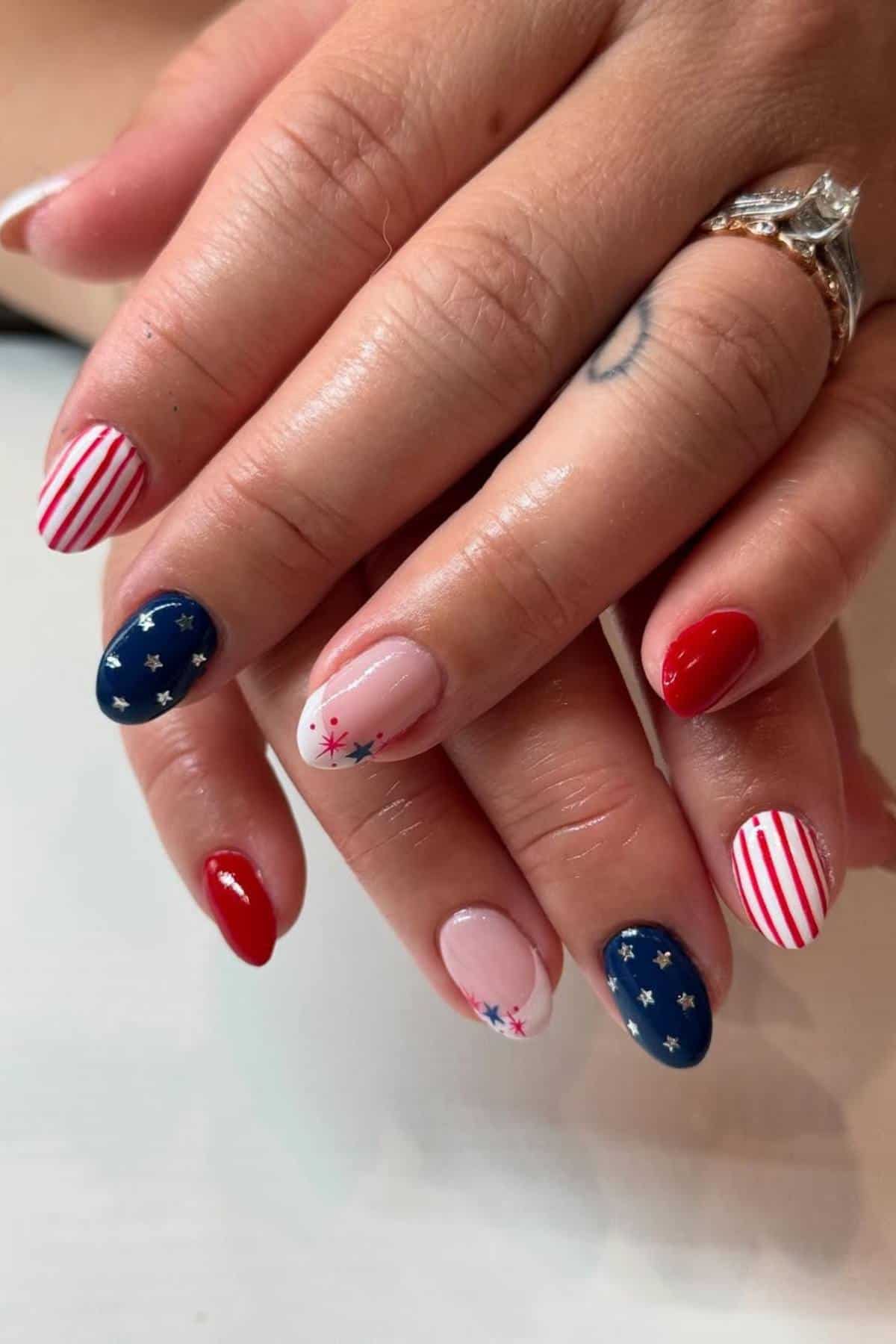 Hands with almond-shaped nails painted in a patriotic theme: red, white, and blue with stripes, stars, and solid colors. A diamond ring is visible on one finger.
