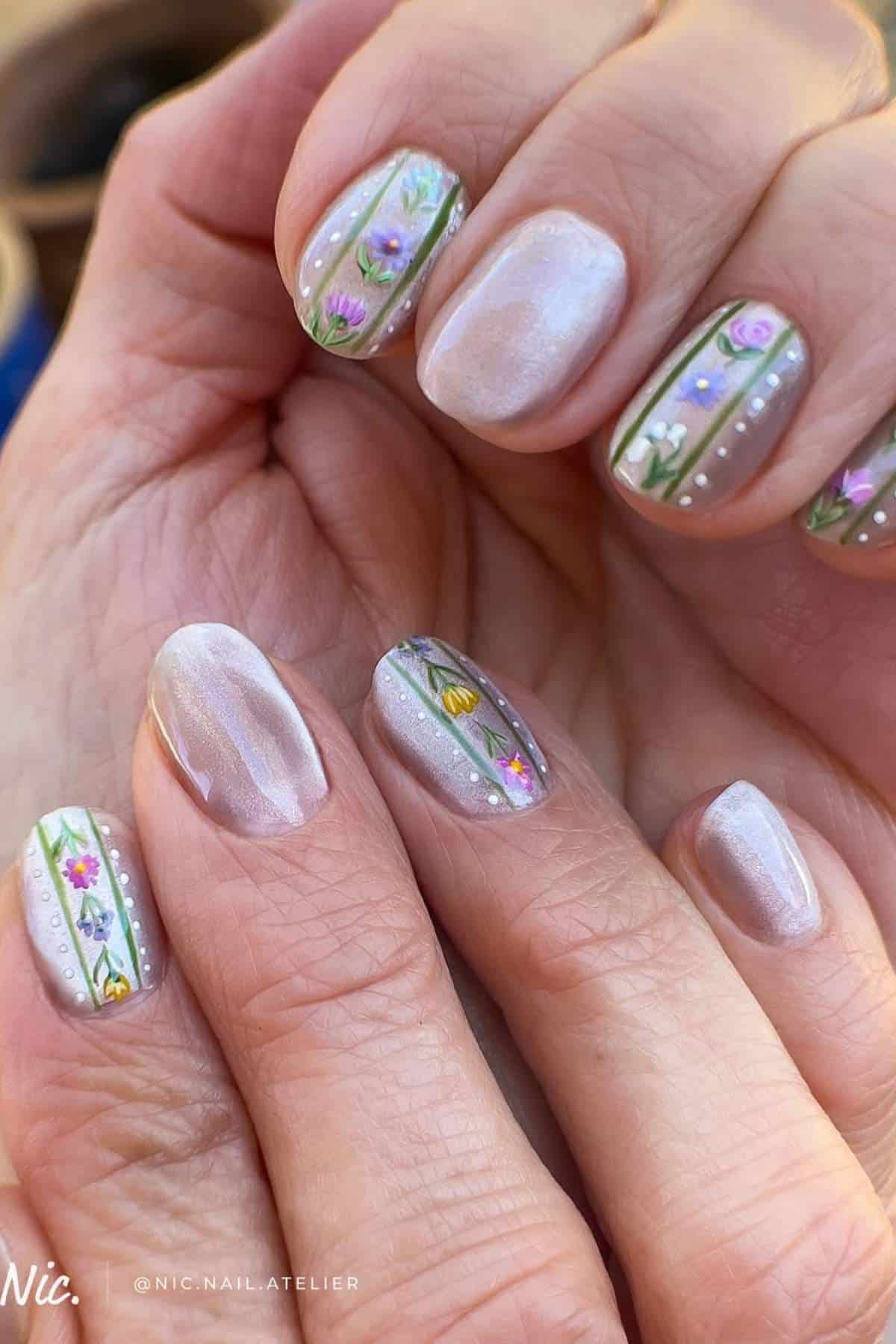 Close-up of hands with light pearlescent nail polish and intricate floral designs on some nails, featuring small colorful flowers and dotted details.