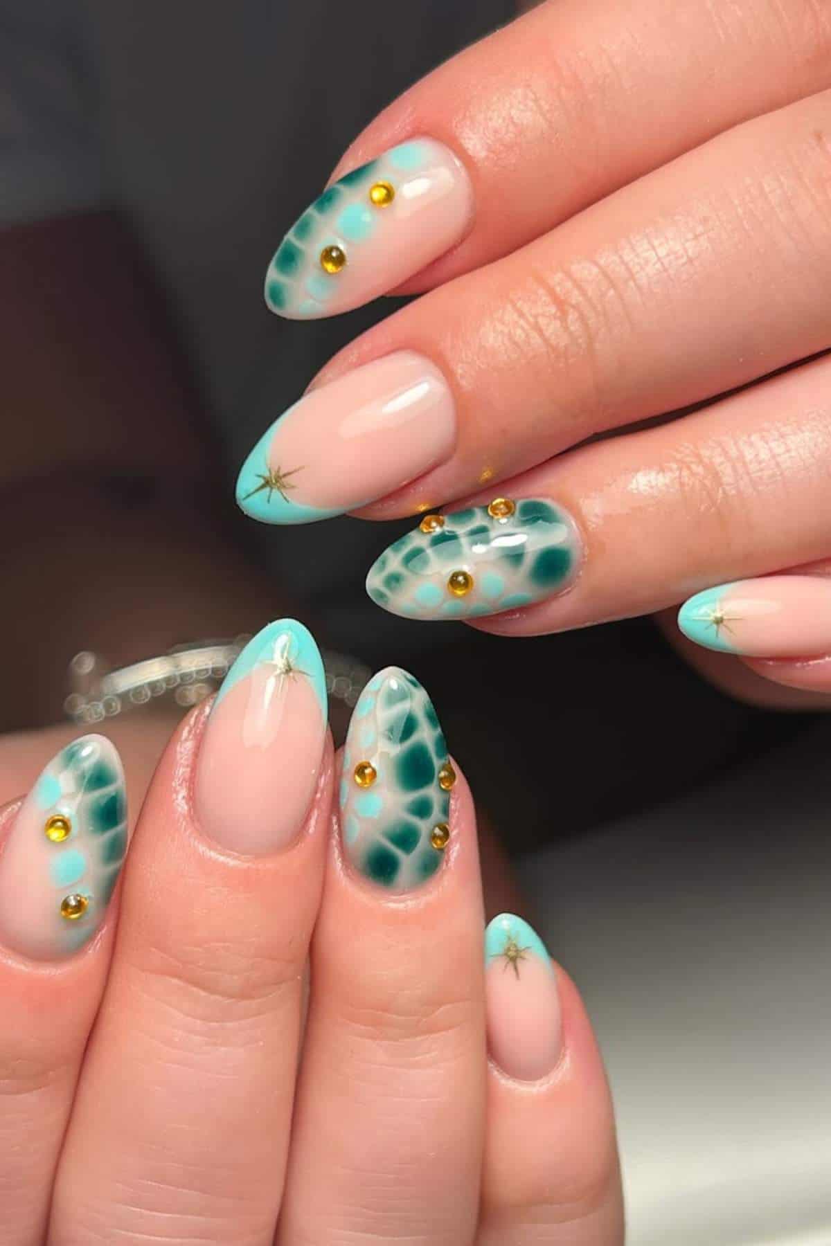 Hands with almond-shaped nails featuring a turquoise and nude geometric design, gold studs, and small star accents.