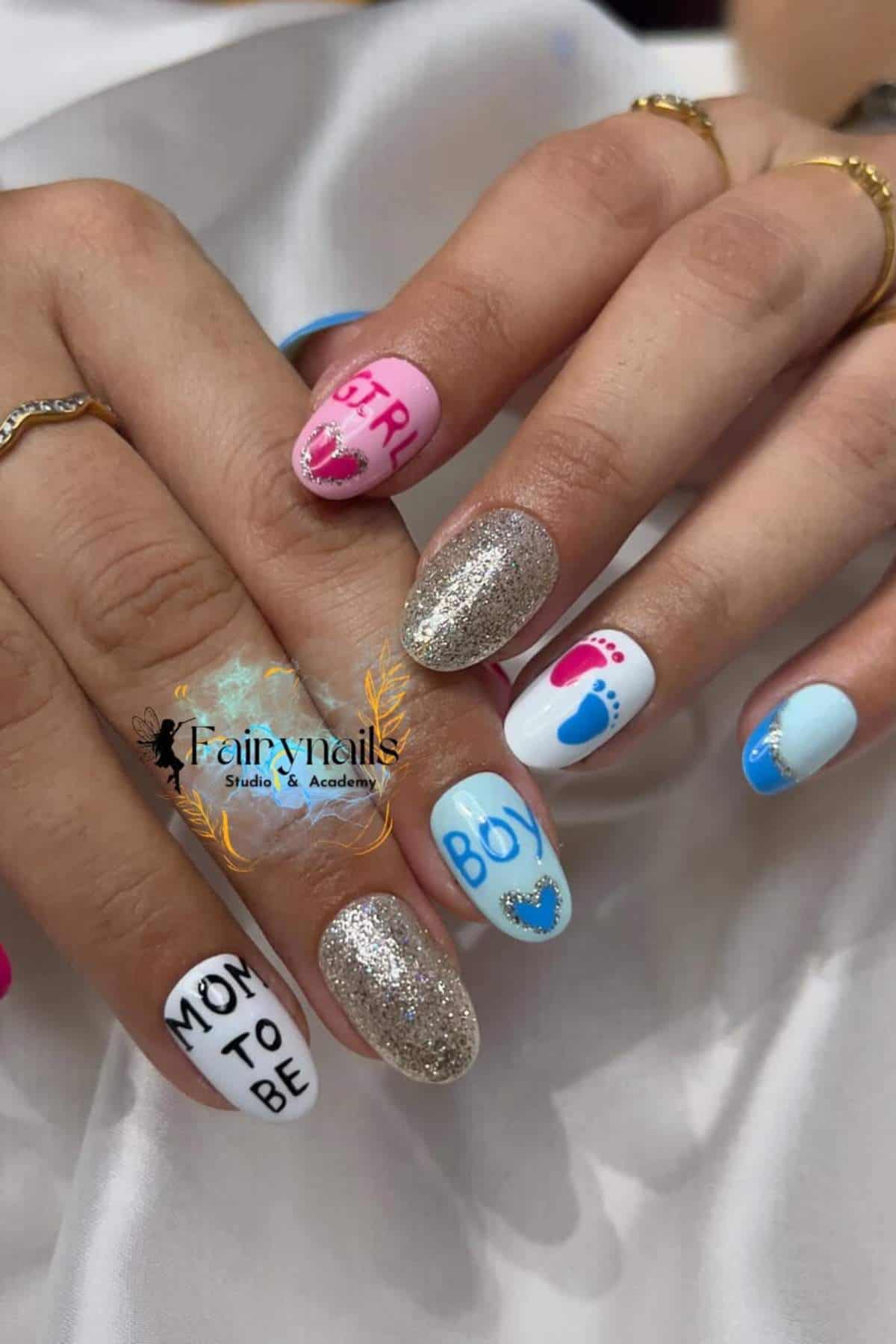 A hand with manicured nails featuring blue and pink designs, glitter, "MOM TO BE," "BOY," "GIRL," tiny footprints, and hearts, suggesting a gender reveal theme.