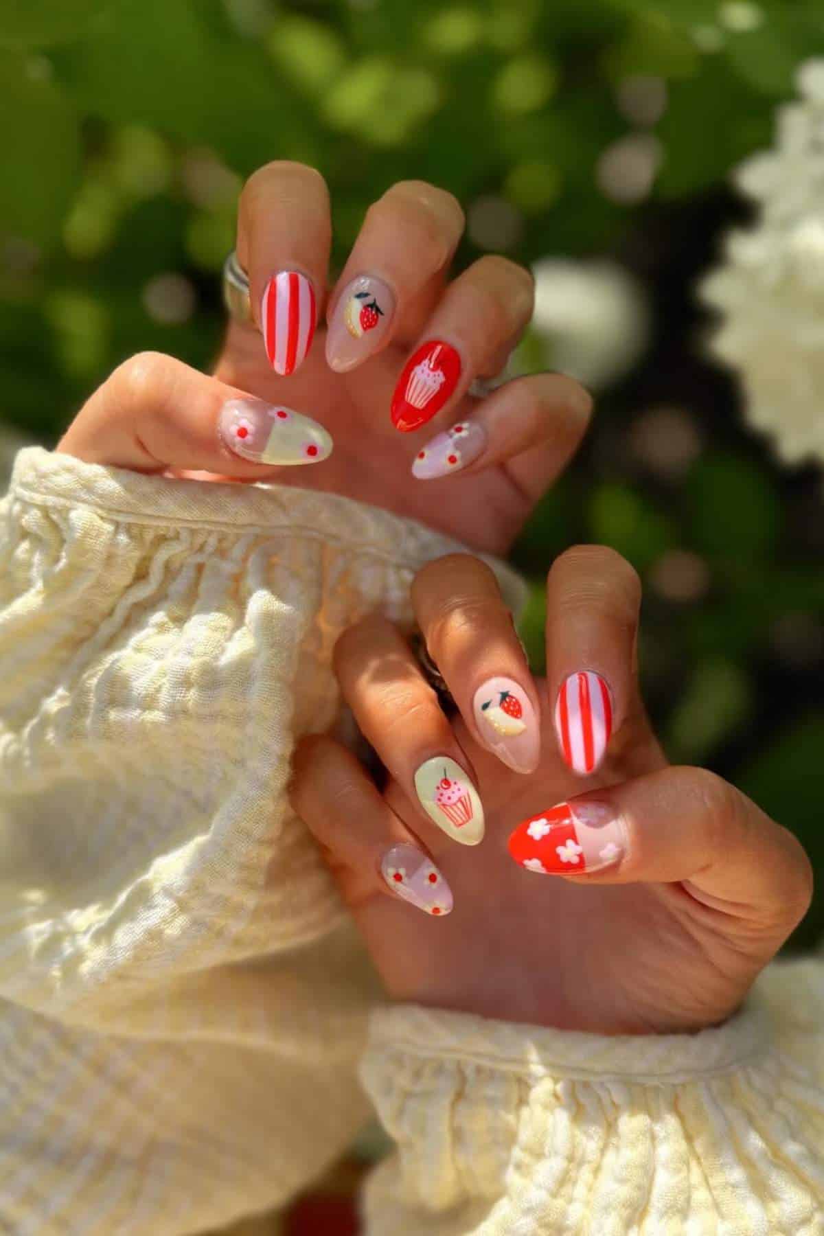 Hands with long, almond-shaped nails featuring red, white, and pink designs, including strawberries, hearts, and stripes, against a blurred green and white background.