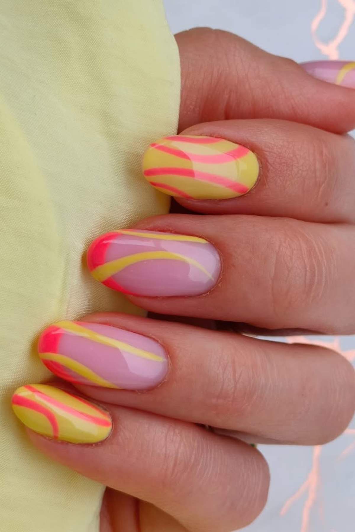 A hand with almond-shaped nails featuring a pastel pink base and abstract yellow and hot pink wavy designs.