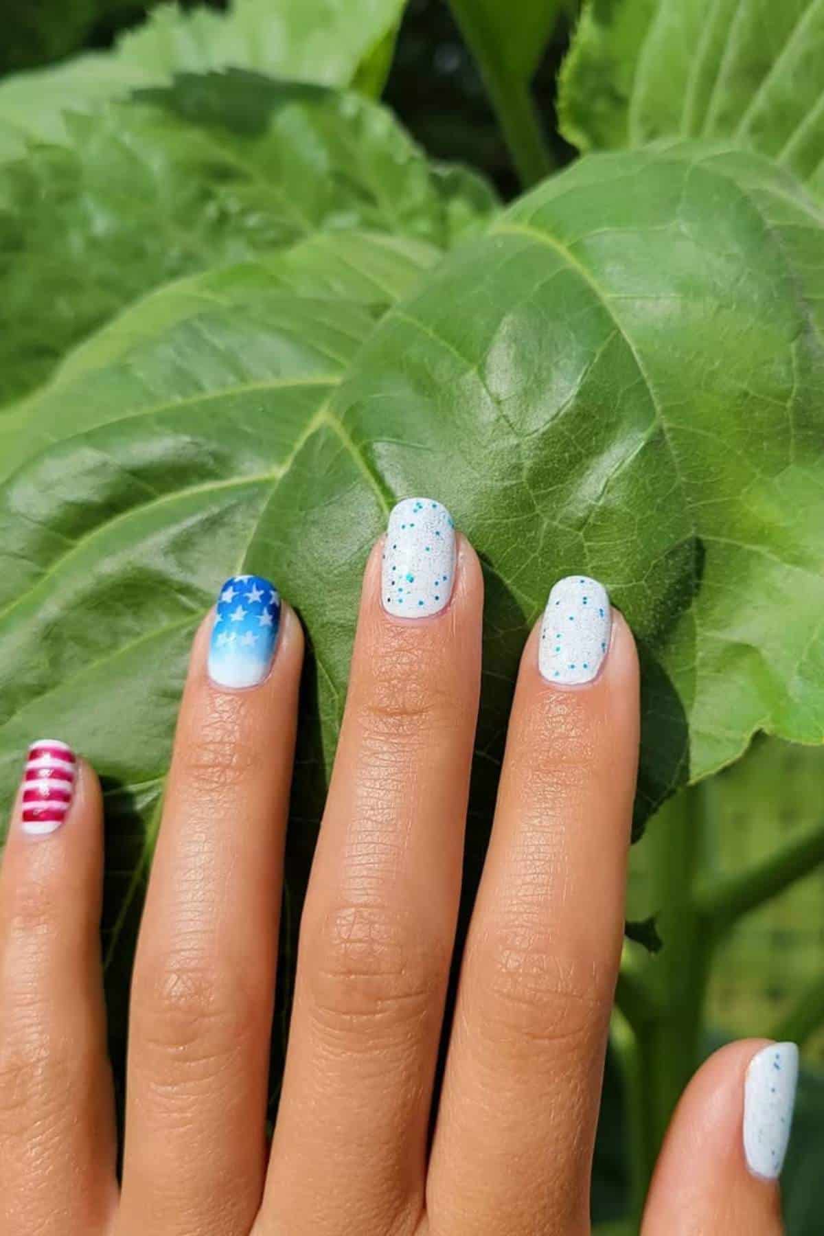 A hand with manicured nails featuring blue and white stars, red and white stripes, and polka dot designs, held in front of large green leaves.