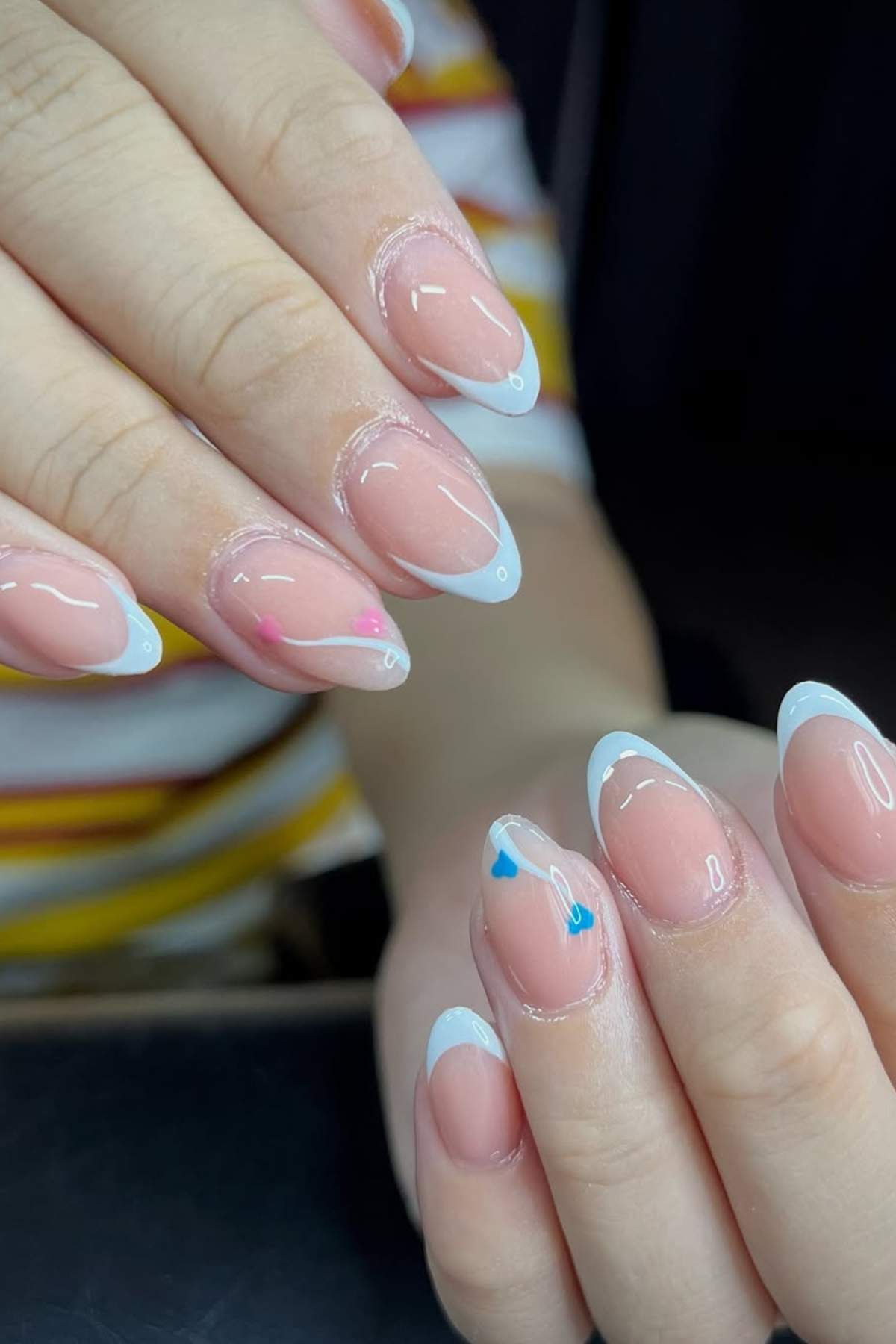 Close-up of hands with almond-shaped nails featuring a natural base, white French tips, and small blue and pink heart designs on the ring fingers.