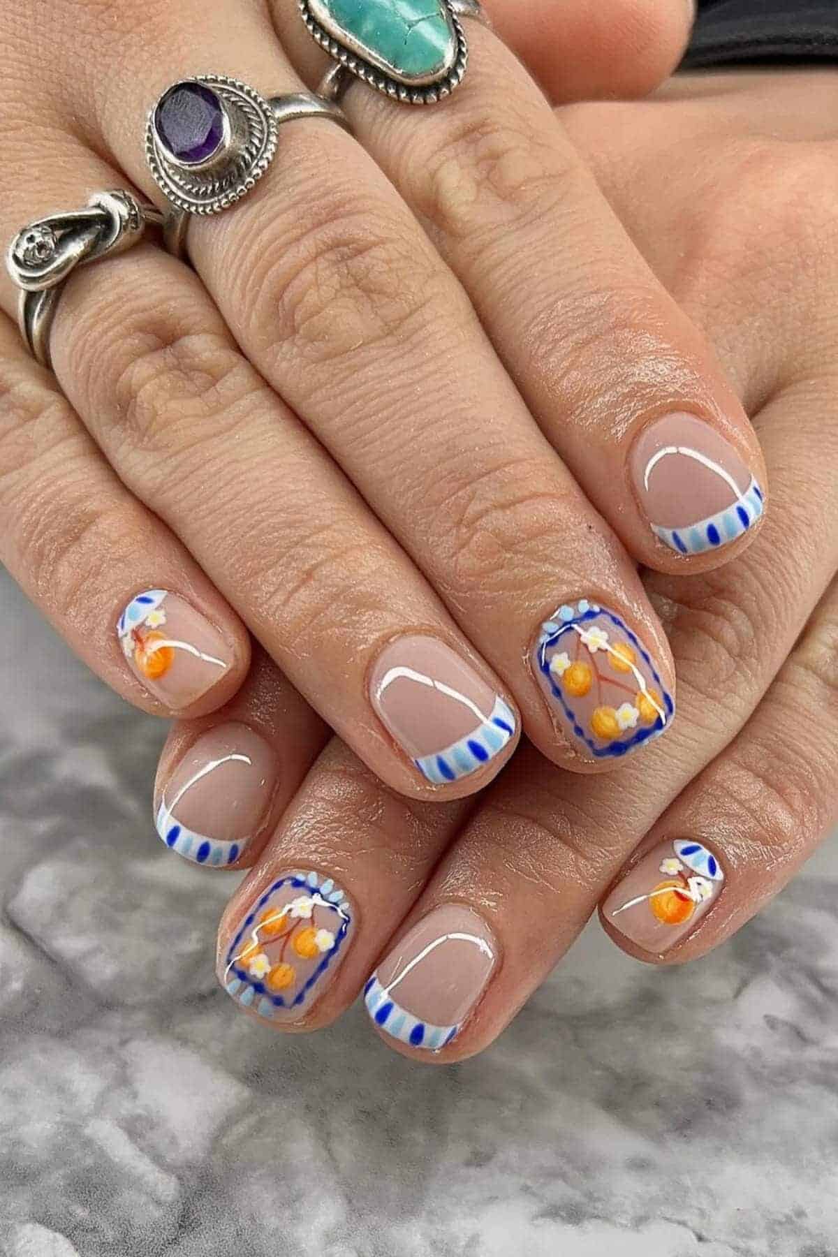 Hands with short, square-shaped nails featuring blue and white striped French tips and orange floral nail art; person is wearing silver rings with turquoise and purple stones.