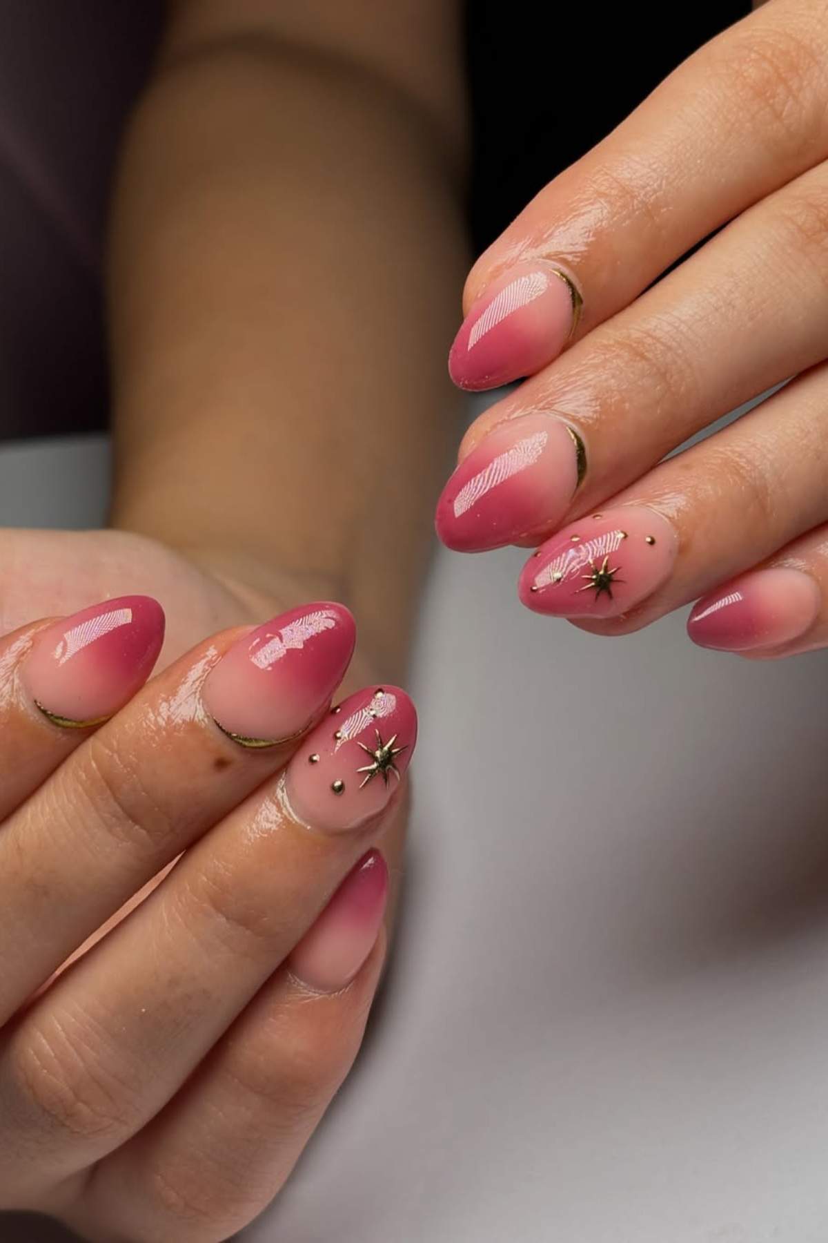 Hands with almond-shaped nails featuring pink ombré polish, small silver star decals, and tiny black dot accents.