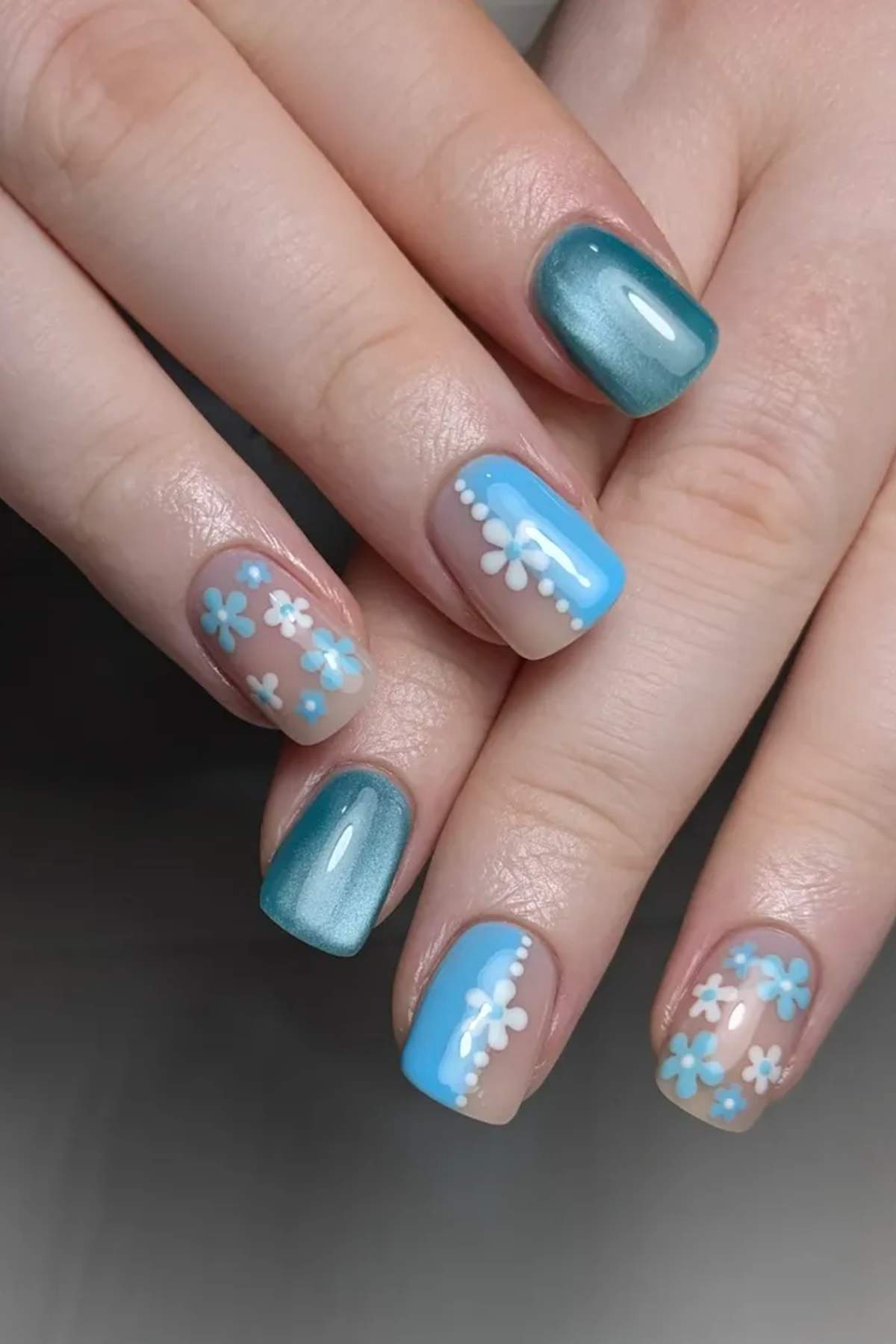 Hands with manicured nails featuring blue polish, metallic accents, and white and blue daisy flower designs on several nails.