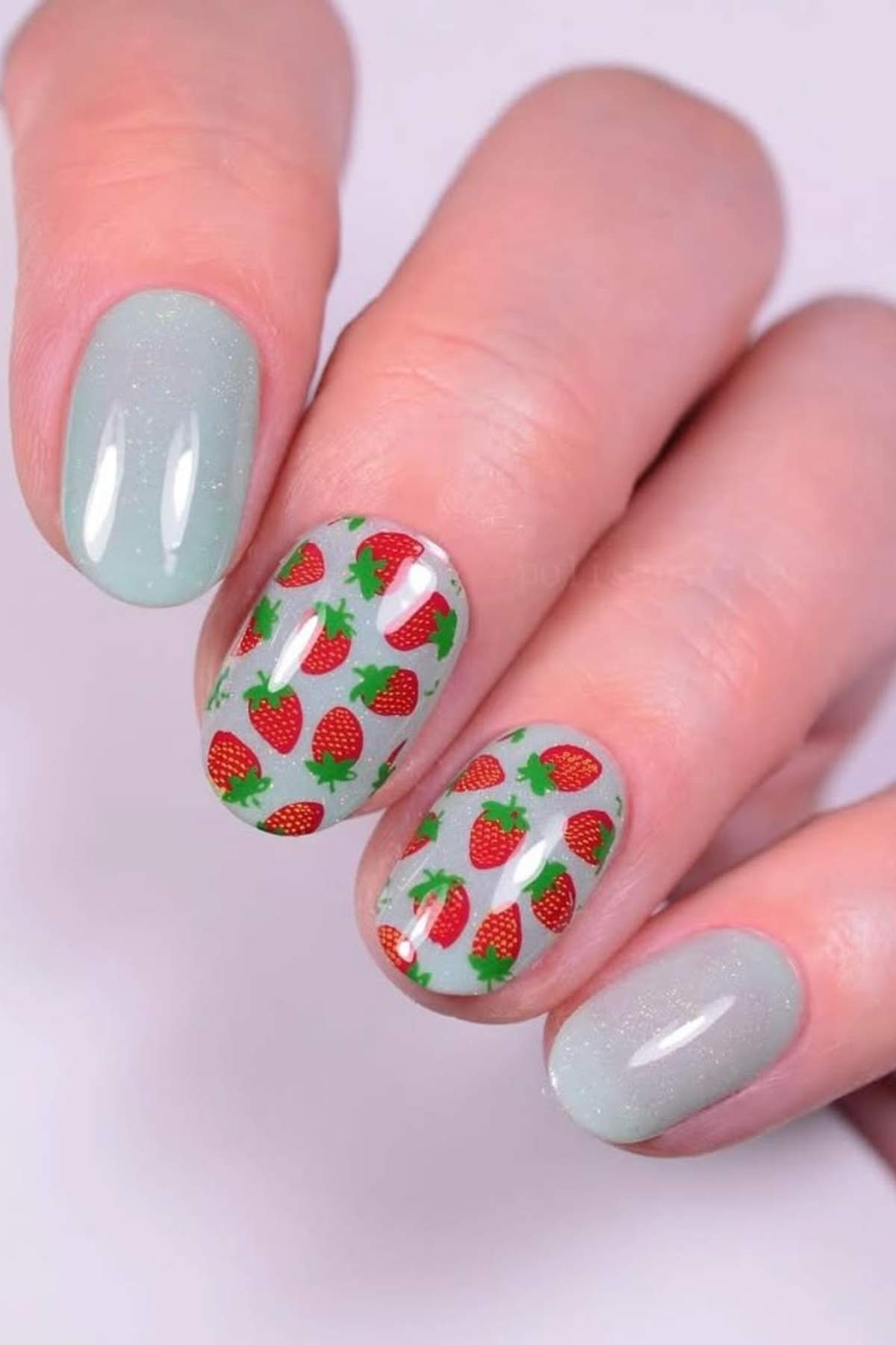 A hand with light blue nail polish on the thumb, pinky, and index finger; the middle and ring fingers have a strawberry pattern on a clear base.
