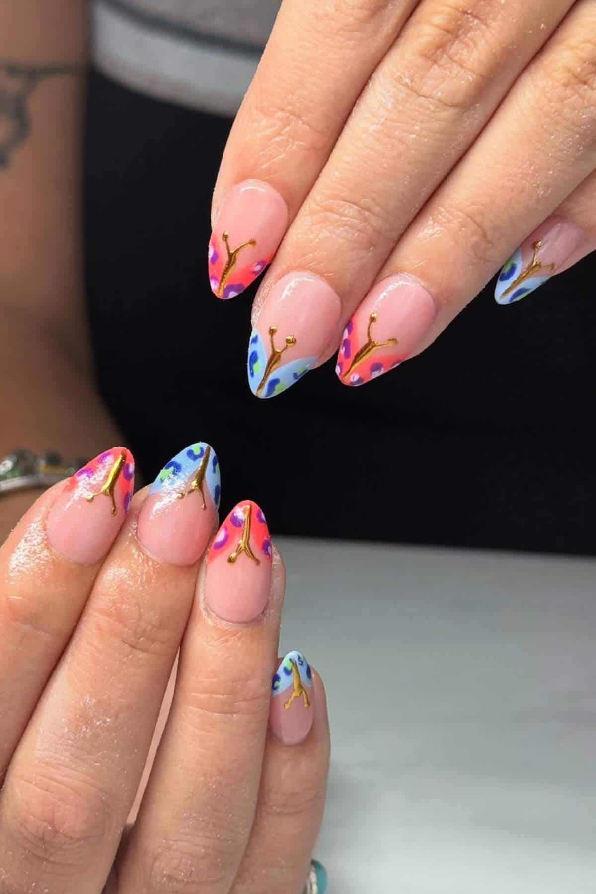Hands with almond-shaped nails featuring pink, blue, and orange abstract designs with gold line accents on a nude base.