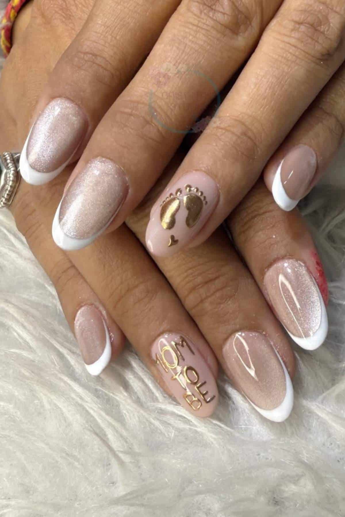 Hands with French manicure on rounded nails, featuring gold baby footprints and "Mom to be" text accent nail, resting on a white textured surface.