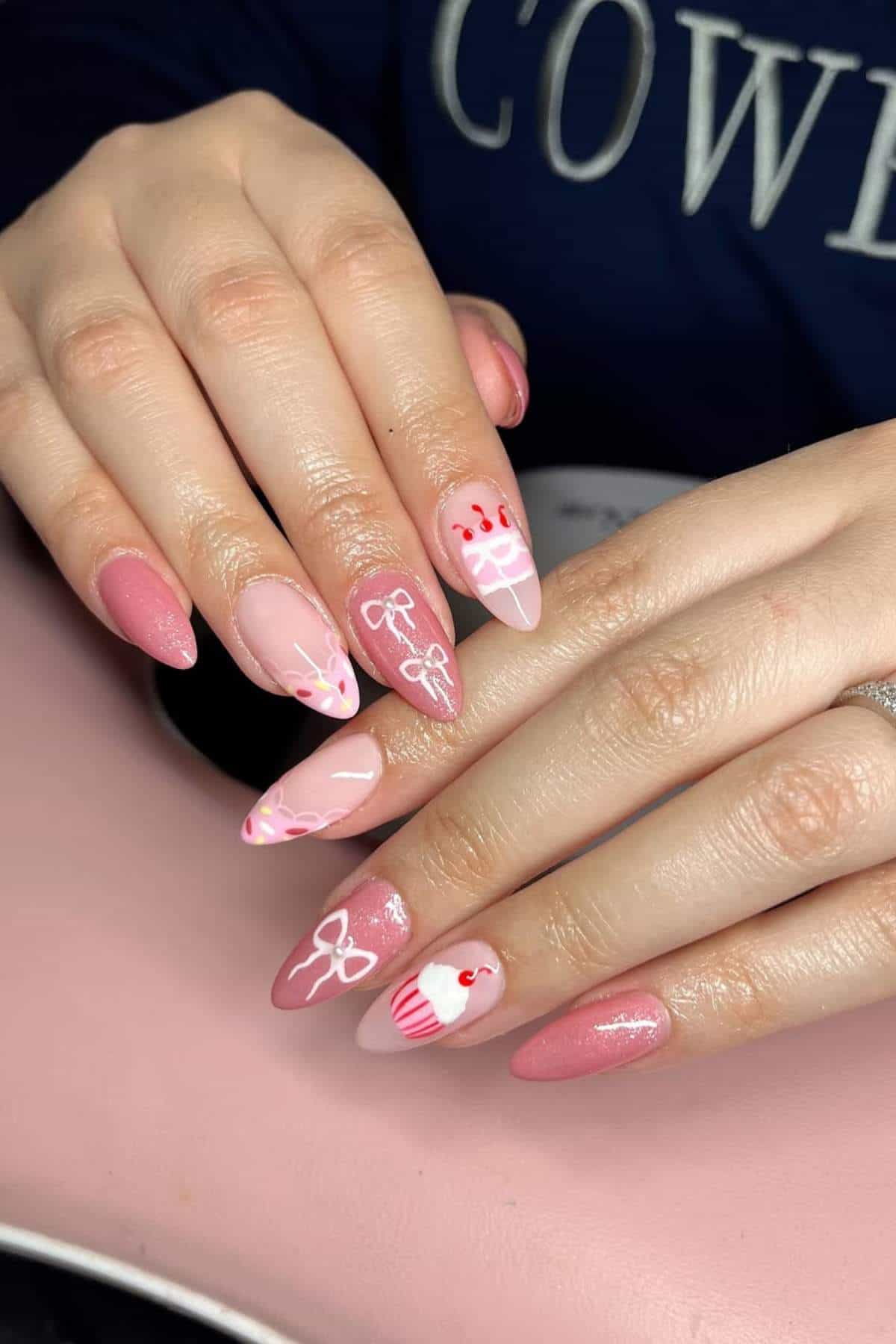 Hands with almond-shaped pink nails, featuring white designs of bows, hearts, and a cupcake on some nails.