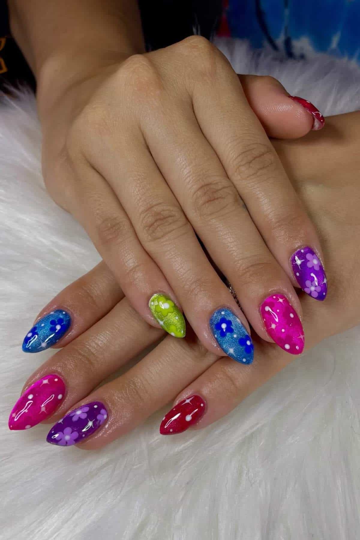 Hands with almond-shaped nails painted in various bright colors, each nail decorated with white flower designs, resting on a white furry surface.