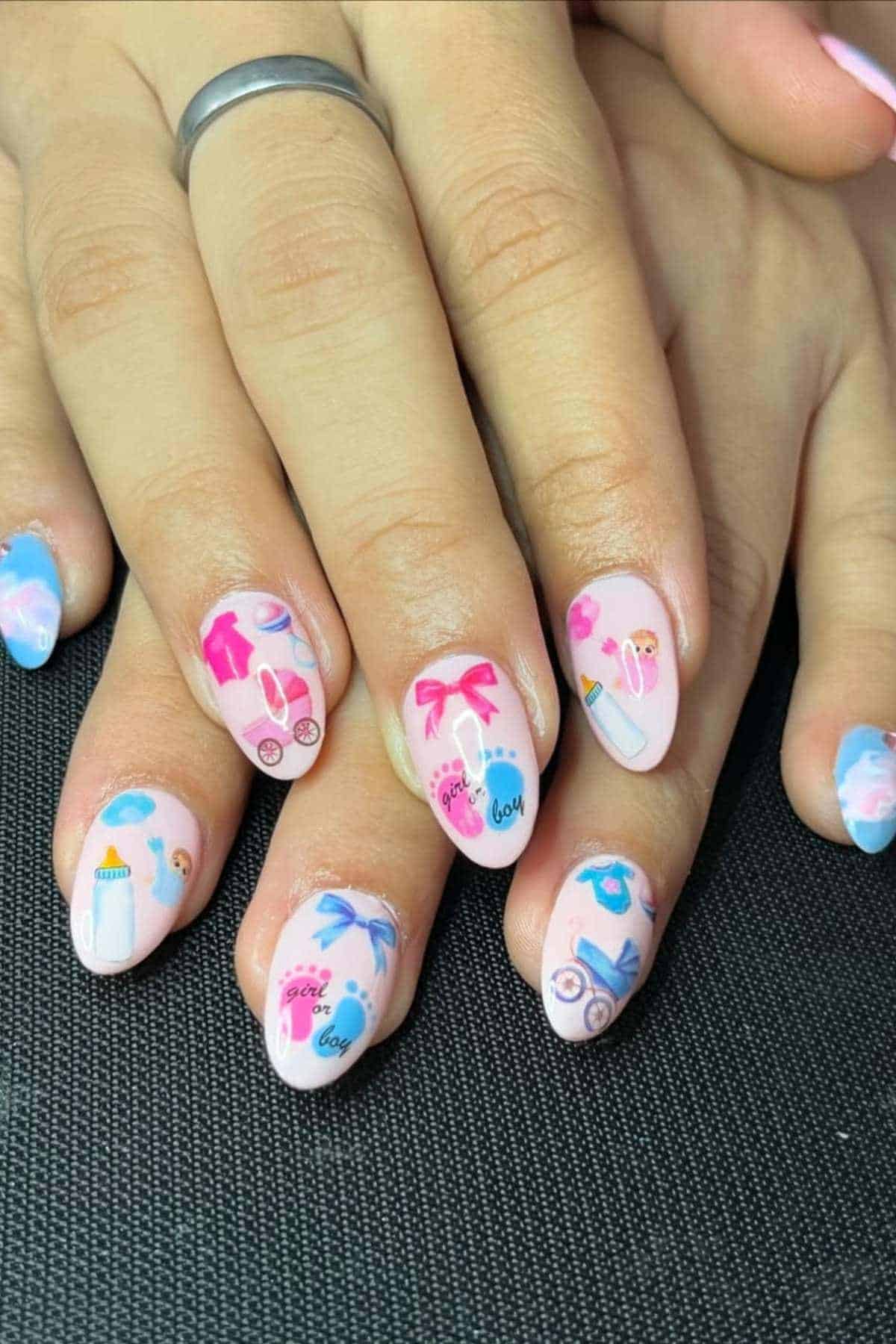 A hand with almond-shaped nails featuring pink and blue baby-themed nail art, including bottles, bows, rattles, and the words "boy or girl.