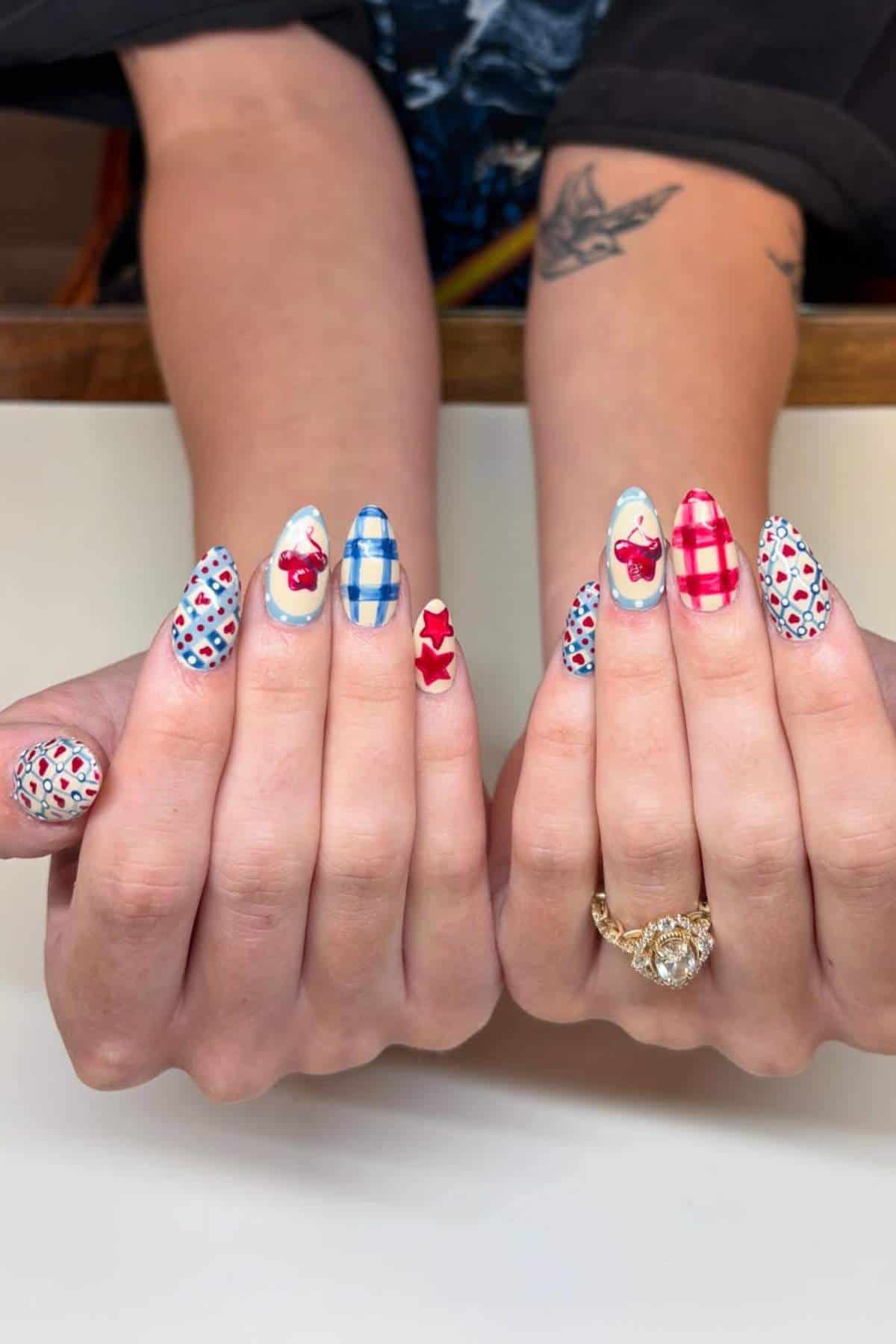 Two hands with long, almond-shaped nails featuring red and blue plaid, cherry, and flower designs. One hand wears a large, oval-shaped ring. A tattoo is visible on one arm.