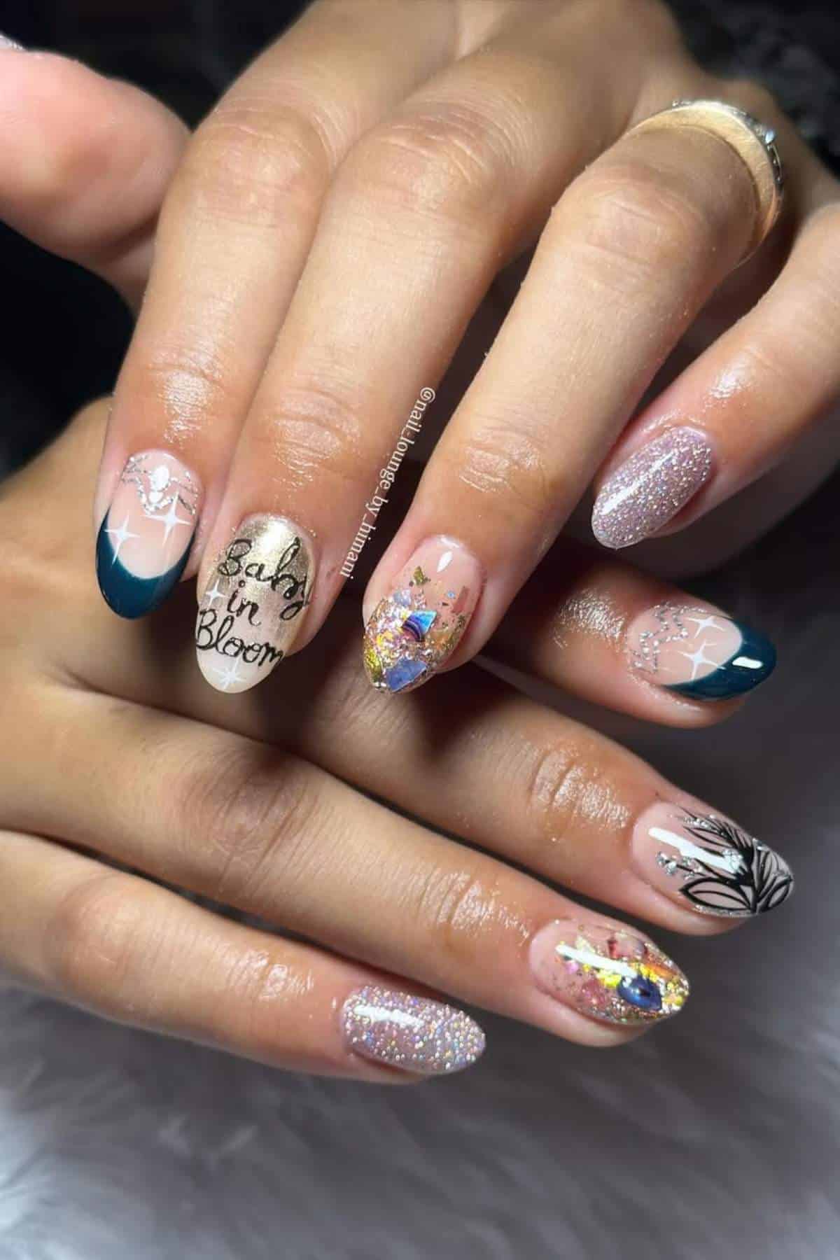 A hand with manicured nails featuring various nail art designs, including glitter, floral patterns, gold foil, and the phrase “Baby in Bloom” on one nail.