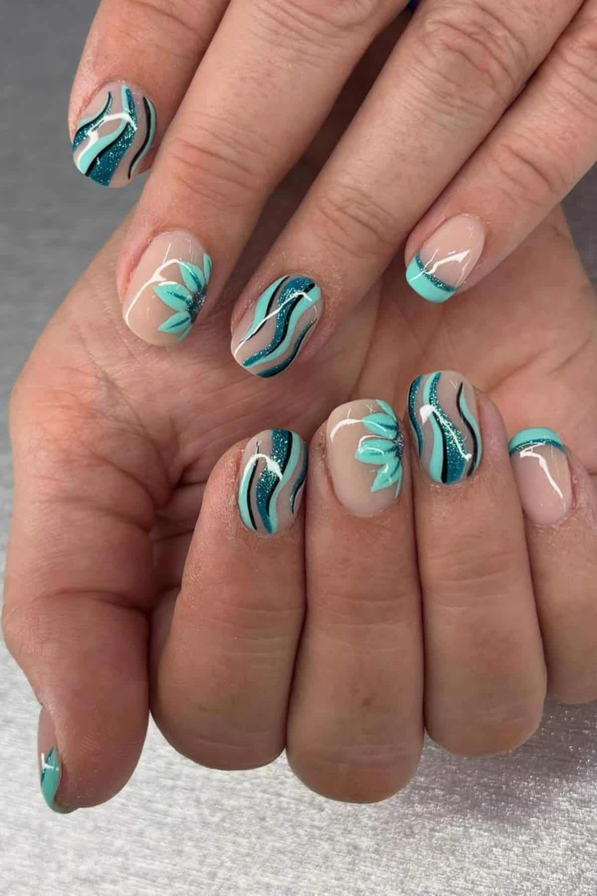 A close-up of hands with nude-colored nails featuring teal, black, and white wavy lines and floral designs on a silver background.