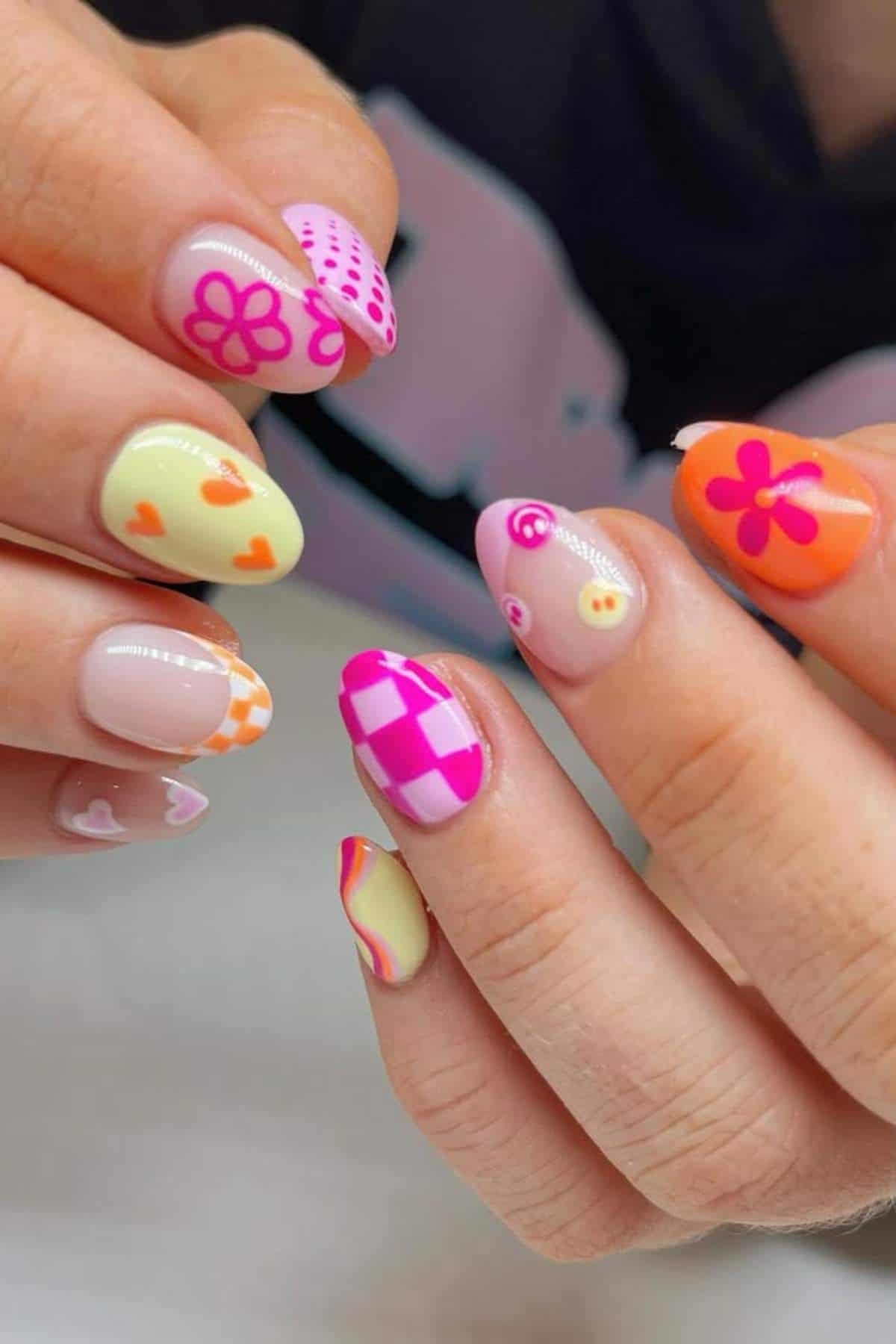 Close-up of hands with almond-shaped nails featuring colorful designs, including flowers, hearts, checkerboard patterns, and smiley faces in pink, orange, yellow, and purple.